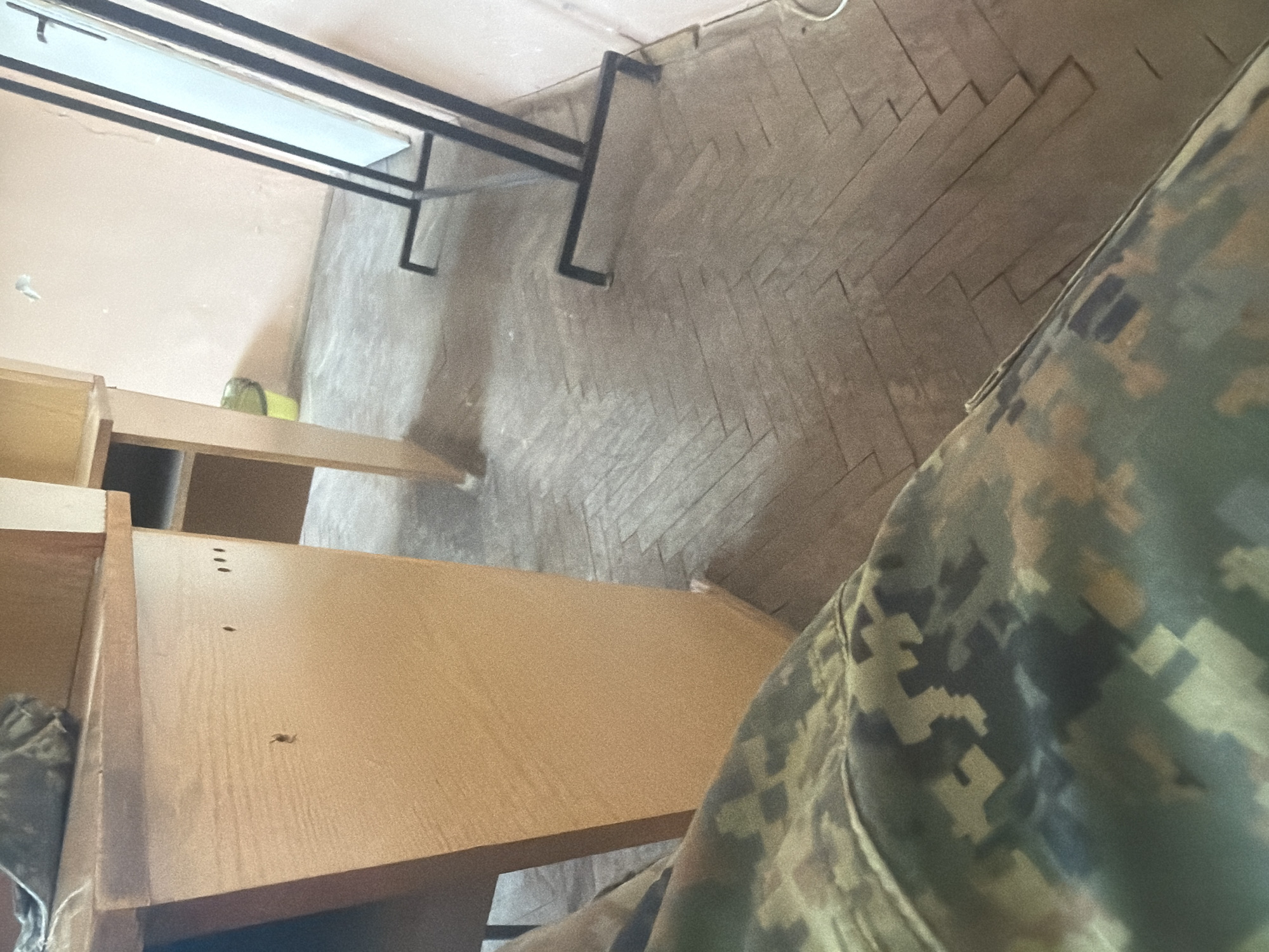 The image shows an indoor scene with a partial view of a person wearing camouflage clothing in the foreground. The background includes a table or desk with a metal frame and a white surface. A wooden structure resembling a cabinet or desk is partially visible on the left side of the frame. The floor is tiled in a herringbone pattern. The location is unknown and no specific details suggest the scene is in Dolna Mitropolia, Bulgaria.