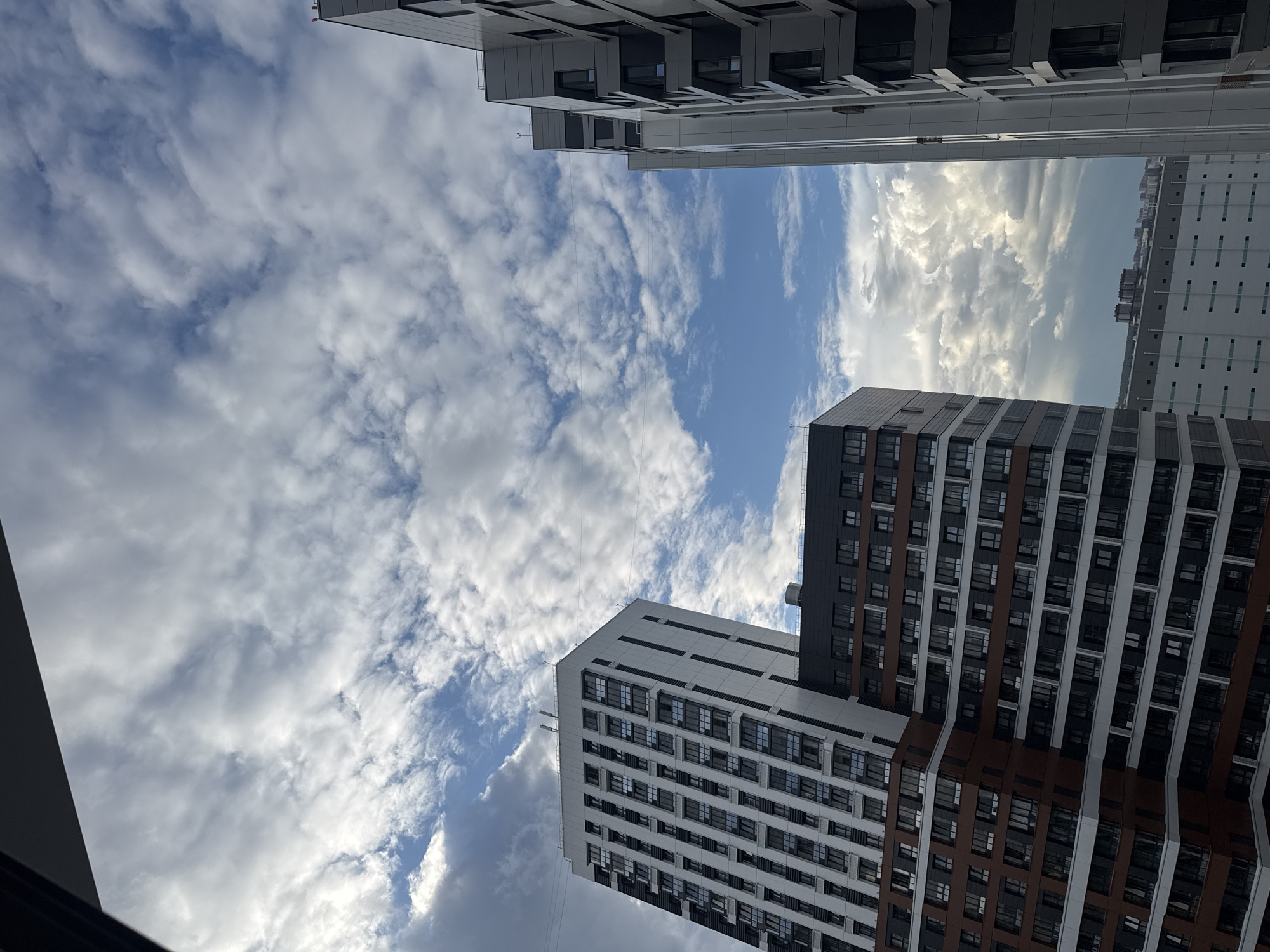 This is a low-angle, medium-shot photograph of a cloudy sky over a residential area in Novosibirsk, Russia. The sky is a mixture of blue sky and white, puffy clouds, with some darker, more ominous clouds in the distance. In the foreground, three modern apartment buildings are visible. The building on the left is tall and white with many windows. The building in the center is shorter and features a dark grey top section and a reddish-brown lower section, also with numerous windows. To the right, another tall, grey apartment building is visible, with a stepped facade. The buildings appear to be under construction or recently completed. The overall scene suggests an urban environment with residential development.