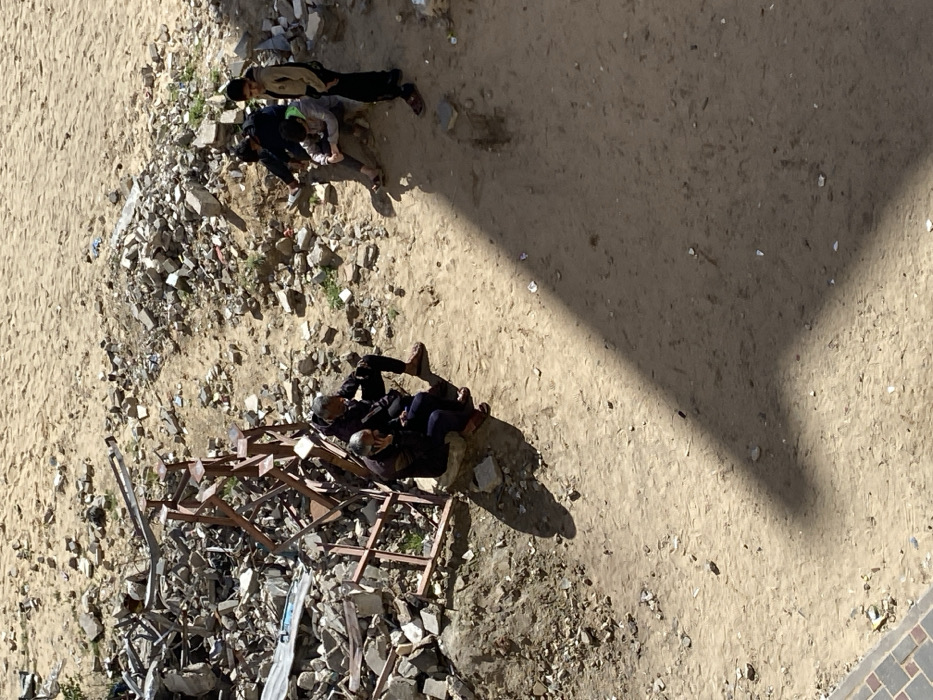 The image depicts an outdoor scene in Khan Yunis, Palestinian Territory, characterized by open ground and debris under bright sunlight. Six individuals are present. On the left, two adult males, dressed in dark clothing, sit on concrete blocks next to a large pile of rubble consisting of broken concrete, metal fragments, and a skeletal metal frame. One of these men holds a mobile phone to his left ear. To the right, four younger individuals, appearing to be males, are grouped near another smaller pile of rubble. Three of these individuals are seated or crouched, while one stands upright, facing right. Strong, distinct shadows are cast across the dirt ground, indicating direct overhead sunlight. The ground is predominantly light brown dirt or sand, with scattered small debris. A narrow strip of paved surface is visible in the bottom left corner.