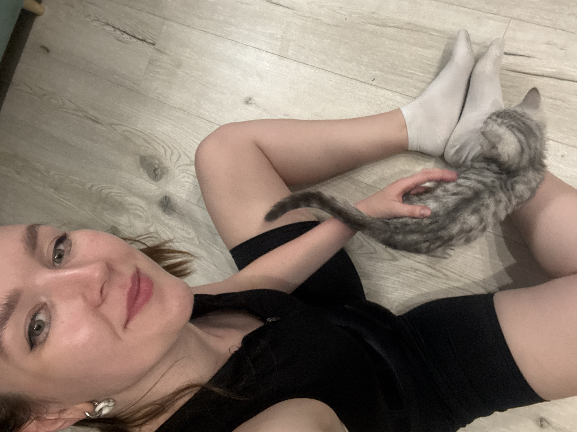 A woman is lying on a wood-patterned floor, looking at the camera. She is wearing a black top and black shorts. She has on white socks. A gray tabby kitten is on her legs and she is petting it with her right hand. The location cannot be determined to be Izhevsk, Russia from the given image.