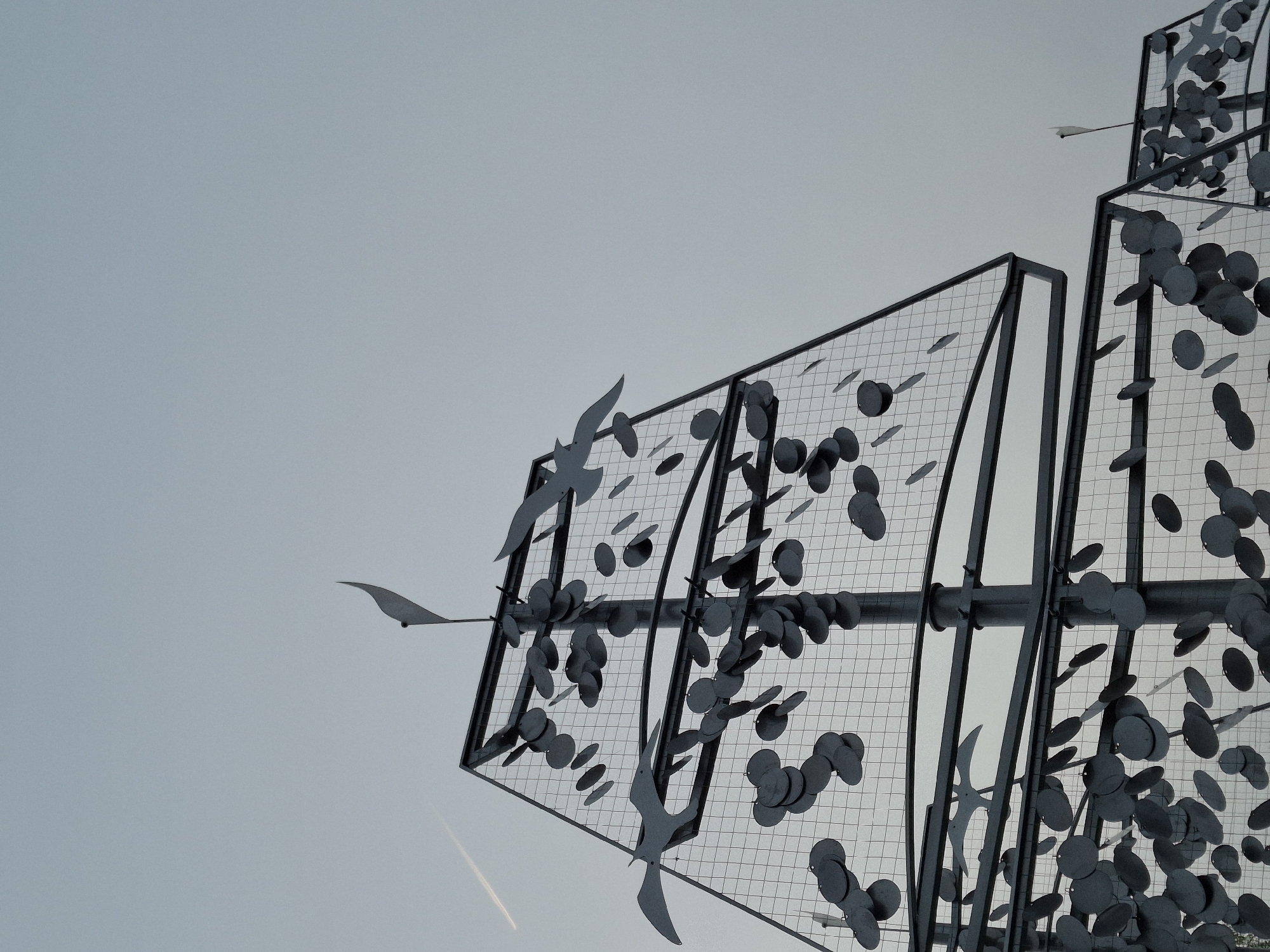A low-angle shot of an outdoor metal sculpture in Chaikovsky, Russia, against a grey sky. The sculpture consists of a rectangular grid framework, partially filled with metallic disks and bird shapes. The sculpture is made up of multiple levels or sections, with the grid pattern extending upwards. The disks and birds seem to be attached to the grid. A contrail is visible in the lower-left area of the sky.