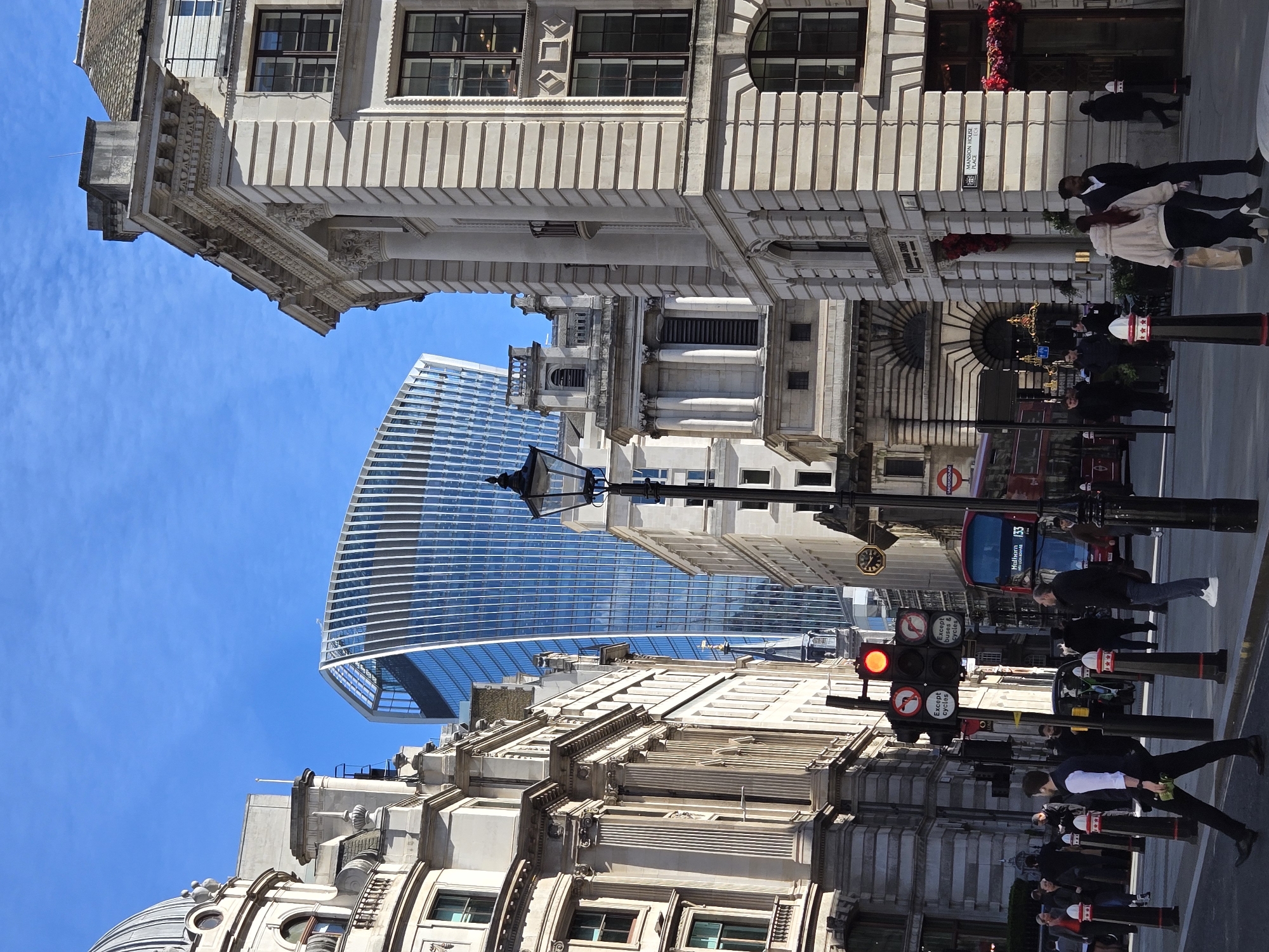 The image depicts a street scene in the City of London, United Kingdom, specifically at the junction of Lombard Street and Mansion House Place EC4, viewed generally southeast. The prominent landmark, 20 Fenchurch Street (The Walkie-Talkie building), a modern skyscraper with a curved glass and steel facade, is visible in the upper-center background. Surrounding it are historic stone buildings featuring classical architectural elements such as fluted columns, ornate cornices, and arched windows.
Approximately 20-30 pedestrians are present on the street and pavements, engaged in walking or waiting. In the lower-left foreground, a male individual in a dark waistcoat and white shirt walks left-to-right, holding green items. On the right, a female individual in a light-colored jacket carries a paper bag.
A centrally positioned traffic light displays a red signal. Attached signage indicates "Except cycles" and "Except buses & cycles." A red double-decker bus, identified as route "133 Holborn," is partially visible behind the traffic light. An "UNDERGROUND" roundel sign is located above the bus. Black ornate lampposts and black bollards with red and white reflective tops are spaced along the street. Street signs clearly indicate "Lombard Street" and "Mansion House Place EC4." The sky is clear blue with sparse white clouds. The scene is illuminated by direct sunlight, casting shadows. An entrance on the rightmost building features red floral decorations.