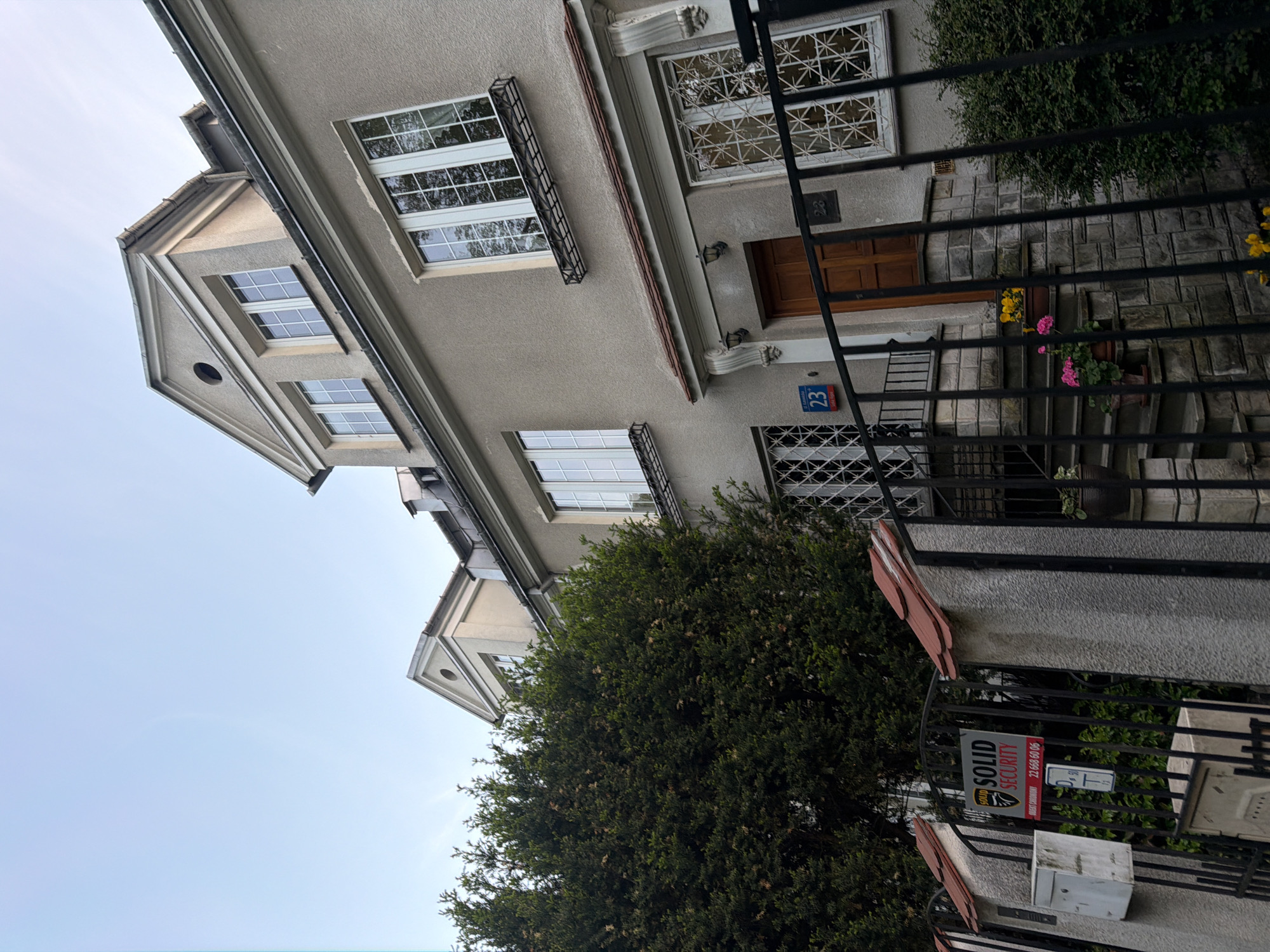 The image shows the exterior of a building with multiple windows and a gable roof. A black metal fence is visible in the foreground, partially obscuring the building's entrance and steps. A "Solid Security" sign is attached to the fence. The building has a "23" address number plate near the entrance. Potted plants are on the steps leading up to the entrance. Trees and bushes partially obscure the facade. The location is Unknown, Unknown.