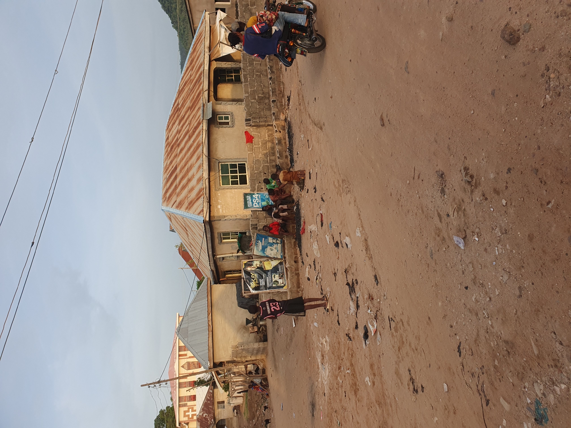 The image shows a street scene in Lokoja, Nigeria. A dirt road runs through the scene, with scattered trash visible on its surface. A group of children are gathered in front of a building, where posters are displayed on the wall. A person wearing a black jersey with the number 23 is standing on the road. A motorcycle with multiple passengers is driving past. The buildings in the background appear to be constructed from concrete, with corrugated metal roofs. Electrical wires are visible overhead.