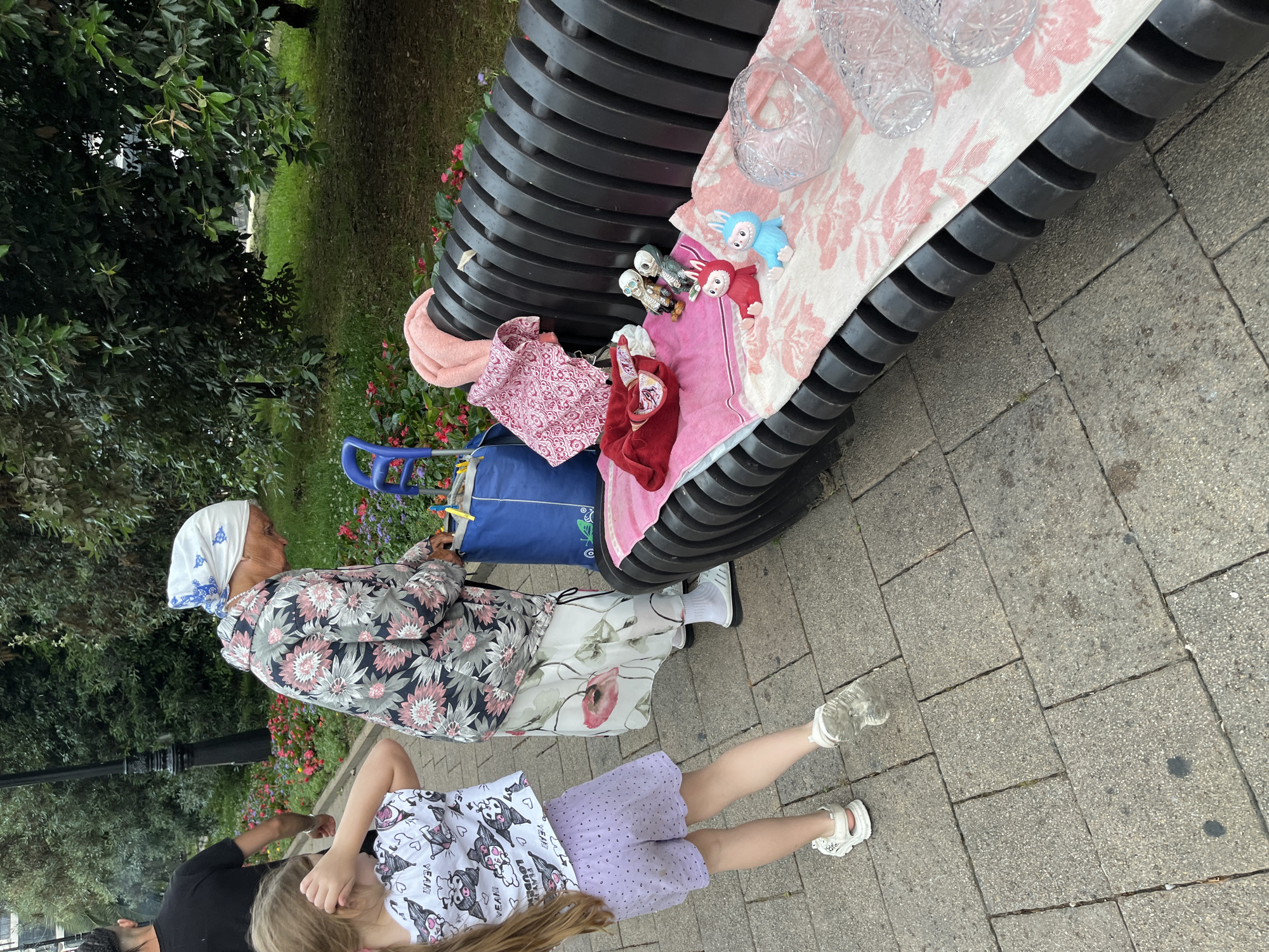 An elderly woman wearing a floral patterned jacket and headscarf is bending over near a blue shopping bag, next to a young girl in a white t-shirt and purple shorts. The girl is facing away from the camera, with her back to the viewer, and her hair is long and light-colored. They are on a paved walkway with a grassy area and trees in the background. To the right, a black park bench is covered with several items, including towels, a pink cloth, a red cloth, two small figurines, and clear glass bowls. The setting appears to be a public park in Sochi, Russia, as indicated by the presence of trees, flowers, and a bench common in urban green spaces. The scene suggests a moment of pause or activity in a park environment.
