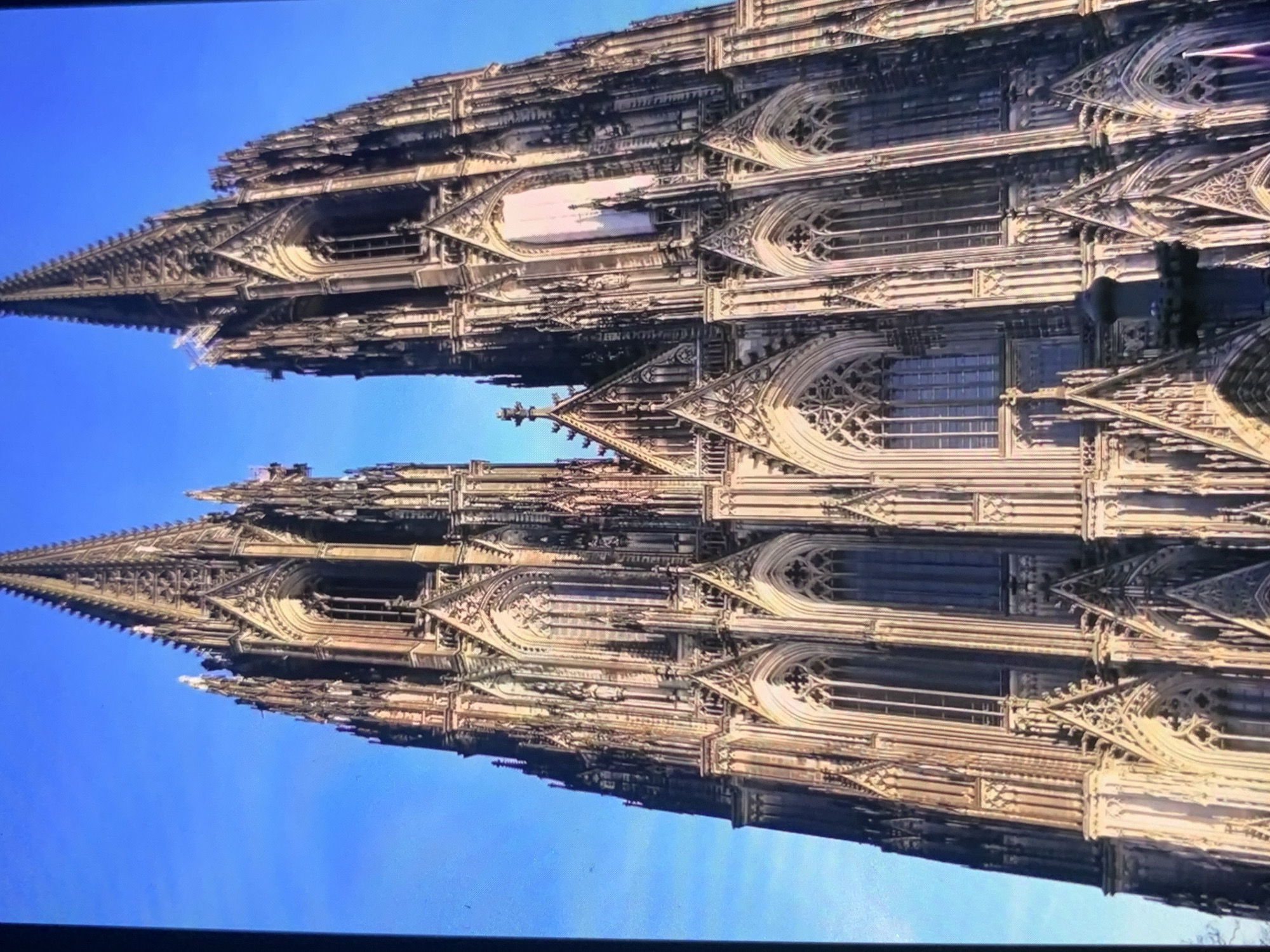 The image is a close-up, low-angle shot of the twin spires of a Gothic cathedral against a clear blue sky. The architecture is highly detailed, featuring intricate carvings, pointed arches, and numerous vertical elements. The stone facade is illuminated by sunlight, highlighting its texture and the shadows cast by the architectural features. There are no people or other objects clearly visible in the frame, except for a dark, columnar structure in the lower right corner, which is partially obscured and could be part of the building's base or an unrelated object. The overall impression is one of grandeur and historical significance, emphasizing the scale and complexity of the cathedral.
This image is not located in Stavropol, Russia. The cathedral depicted is Cologne Cathedral, a UNESCO World Heritage site located in Cologne, Germany.