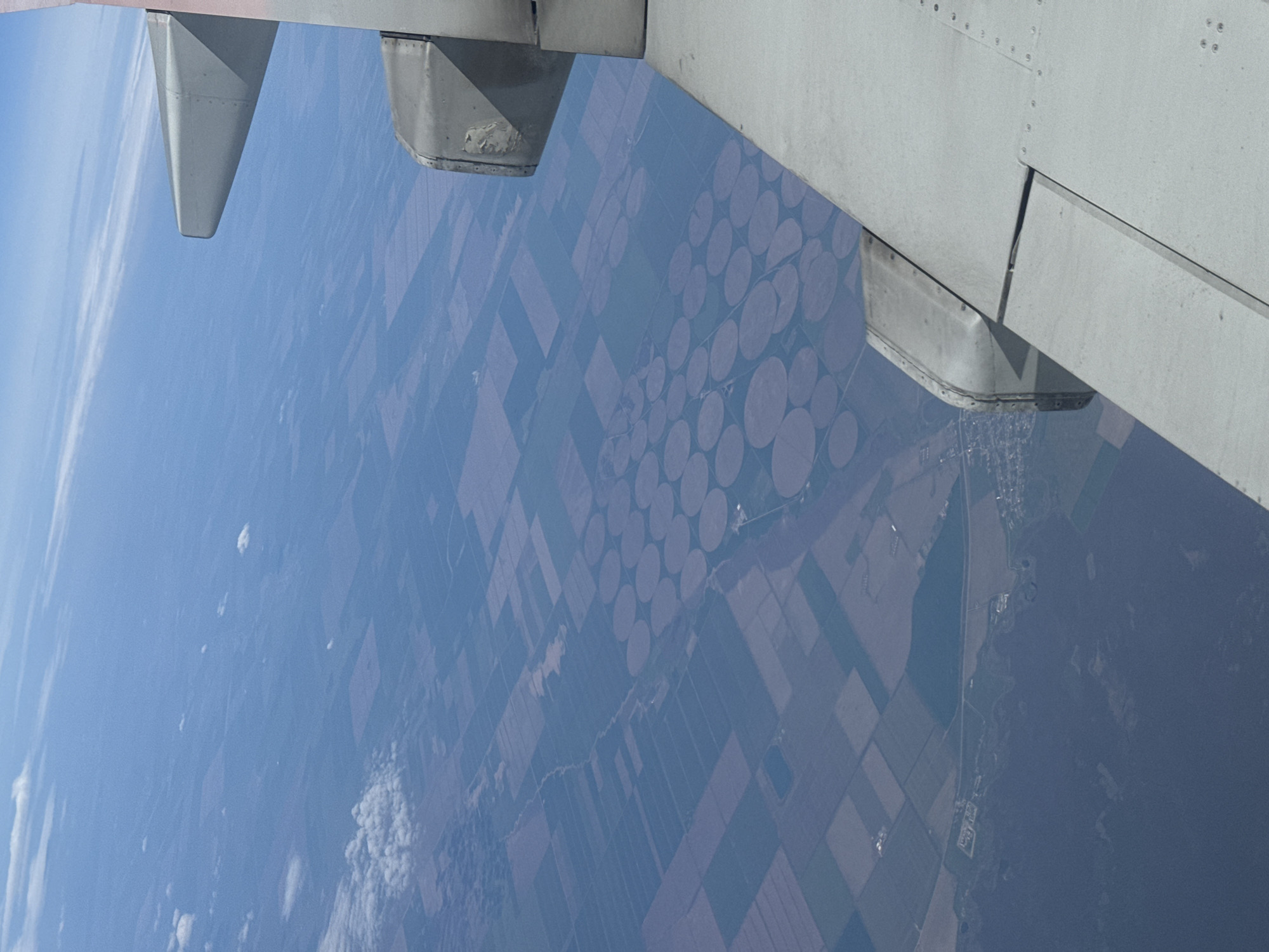 The image is an aerial view from an airplane window, showing land below. Part of the airplane's wing is visible in the upper portion of the image. The land below consists of a patchwork of rectangular and circular agricultural fields. Some of the circular fields are clustered together. A small river or stream meanders through the agricultural area. A settlement is visible in the lower right corner of the image. The scene appears to be in a rural area. The location is potentially Забегалово, Russia, according to the prompt. There are no people visible in the image.