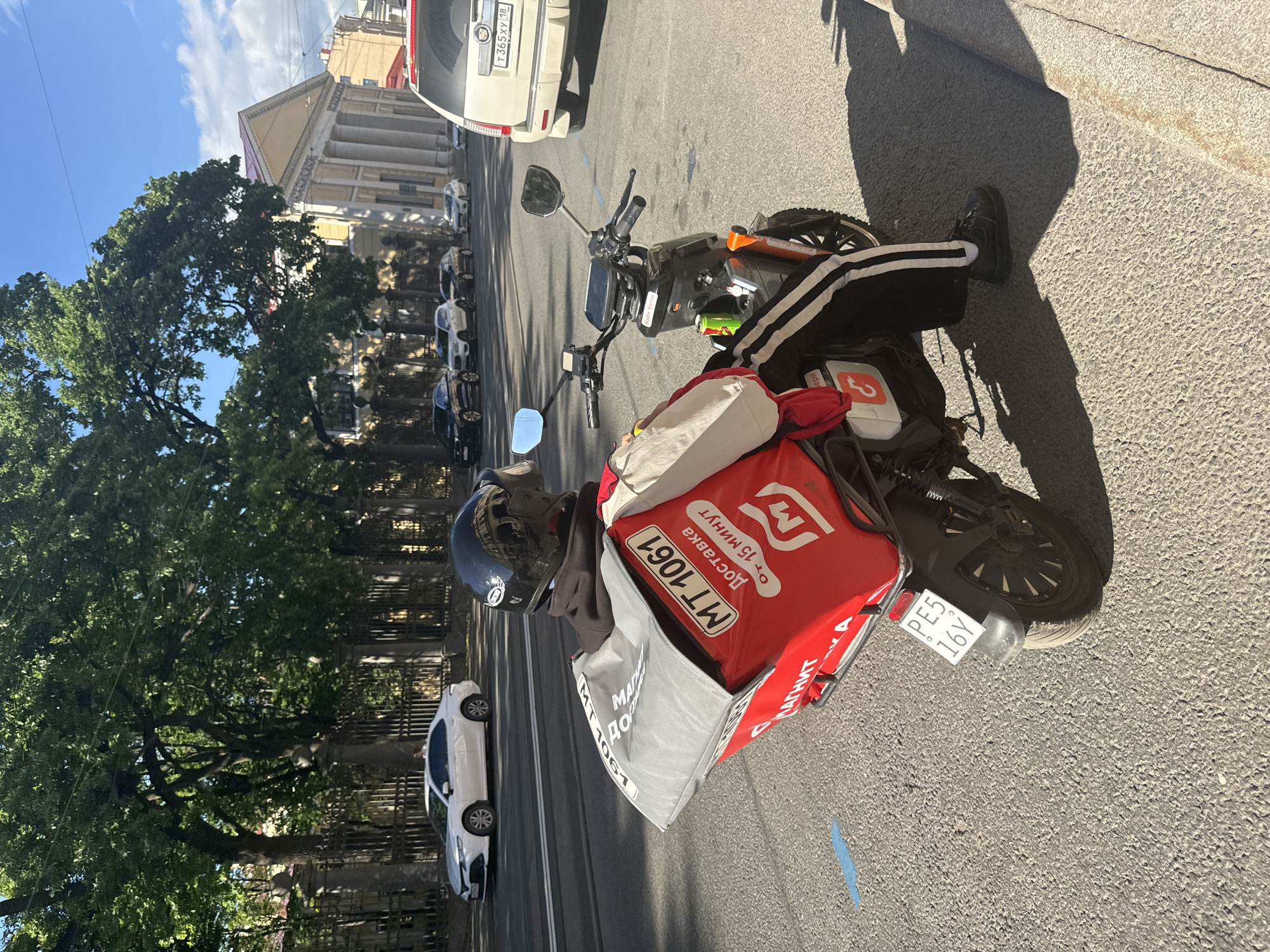 A delivery person on a scooter is parked on the right side of a street in Saint Petersburg, Russia. The delivery person is wearing a black helmet and a dark hoodie with a red and white delivery bag attached to the back of the scooter. The bag has Cyrillic text, including "MT 1061," "Доставка," and "ОТ 15 МИНУТ," indicating a delivery service promising delivery within 15 minutes. The scooter has a license plate that reads "PE5 16Y." In the background, there are trees, buildings with classical architecture, and several cars driving on the street, including a white sedan and a cream-colored SUV. The sun is shining, casting shadows on the street.