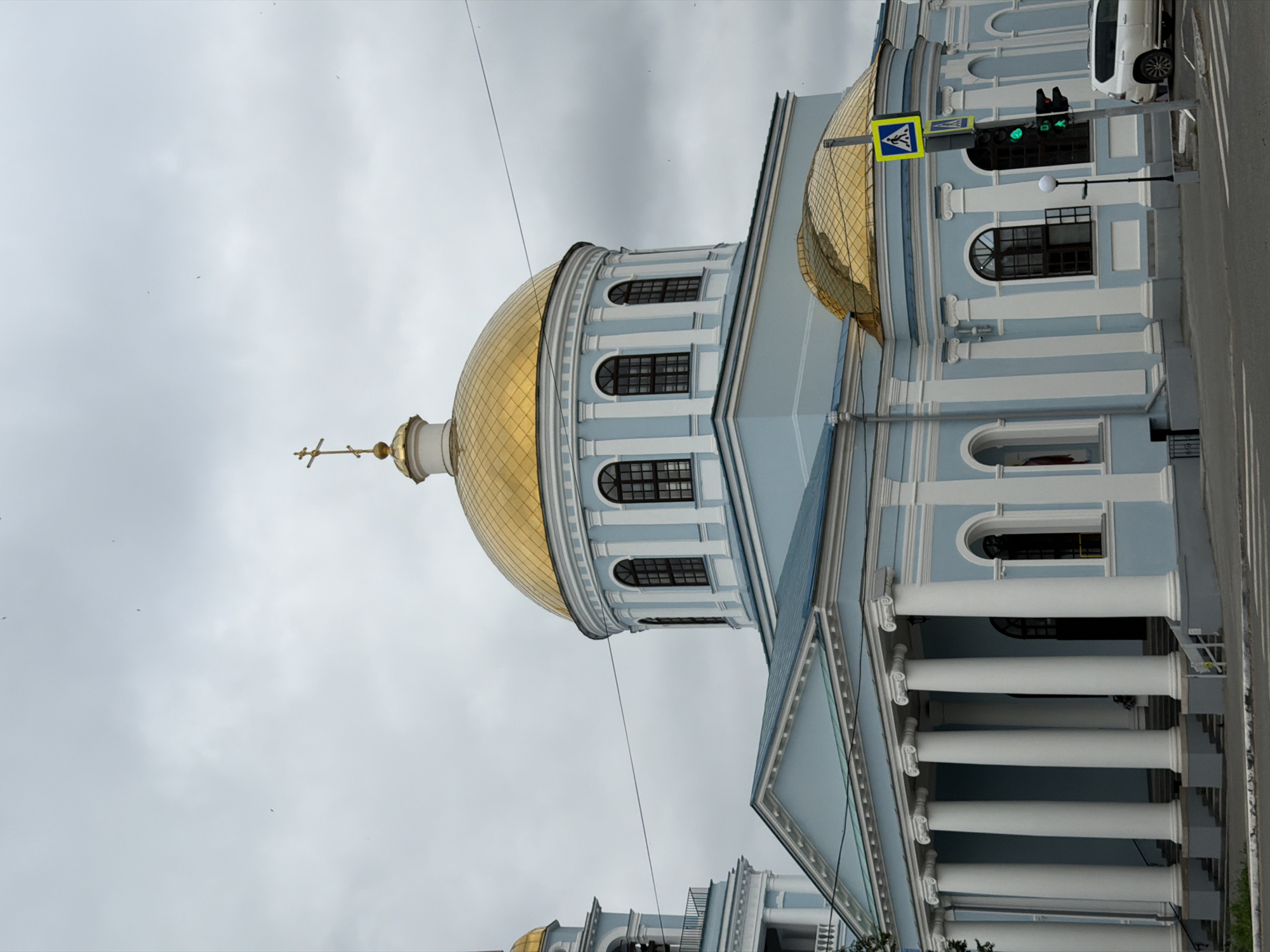 The image shows a bright blue Orthodox church with a golden dome, located in the city of Votkinsk, Russia. The church has a classical architectural style, featuring white columns, arched windows, and decorative moldings. In the foreground, a street with a crosswalk is visible, along with a traffic light showing a green signal for pedestrians. A white SUV is parked on the right side of the image, and power lines run across the sky. The sky is overcast with clouds.