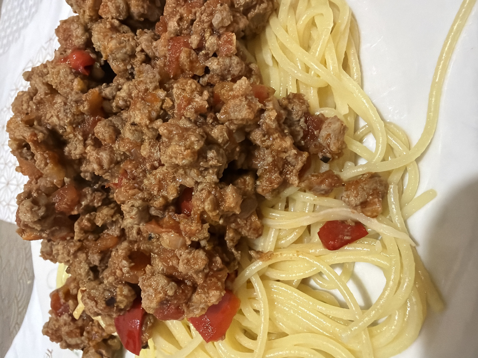 A close-up shot shows a plate of spaghetti with meat sauce. The spaghetti is a light yellow color and arranged in a somewhat circular pile on the right side of the plate. The meat sauce is on the left side, a thick mixture of ground meat, tomatoes, and other ingredients, and is a brownish-red color. The scene is a domestic setting, and there are no apparent indications that would suggest a specific location.