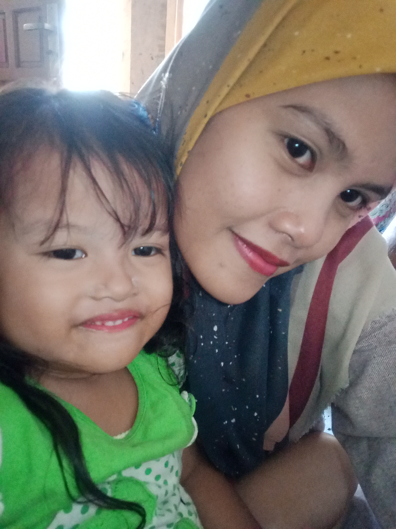 The image shows a selfie of two females in an indoor setting. On the left is a young girl with dark hair, wearing a green shirt with white polka dots. She is smiling. On the right is an adult female wearing a yellow headscarf and a colorful garment. She is also smiling. The background appears to be a room with a wooden door and a window with bright light. The overall impression is a casual and personal moment captured in a residential environment. The location of Juwana, Indonesia, is not discernable from the visual information present in the image.