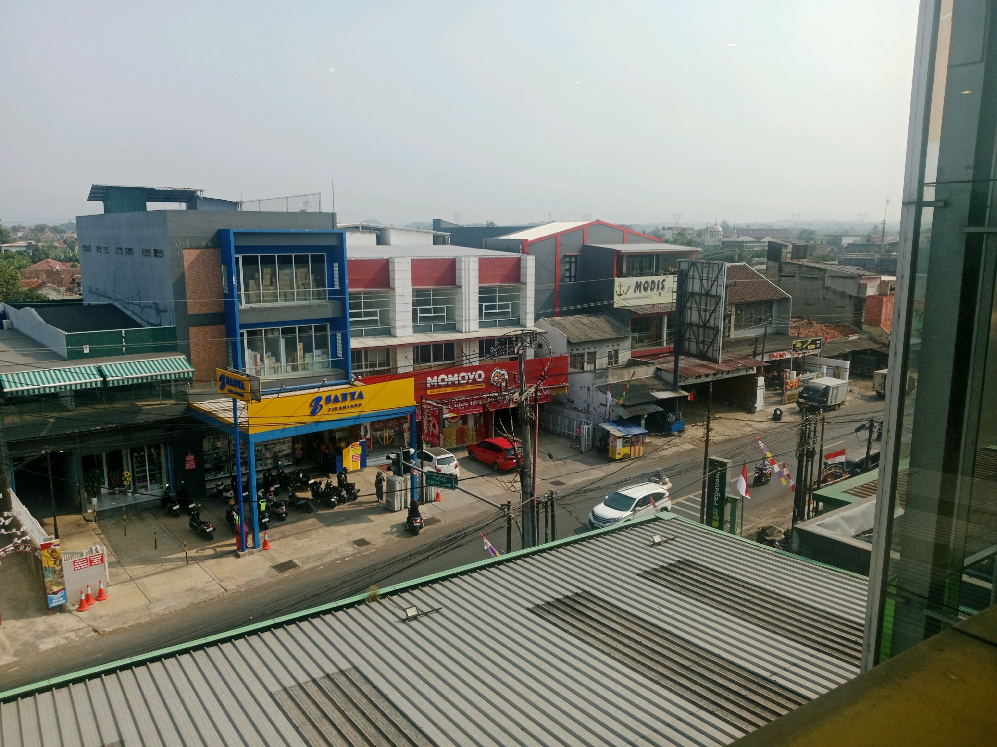 The aerial view captures a street scene in Ciranjang, Indonesia, featuring a row of commercial buildings. On the left, a blue and grey building with large glass windows is visible. Adjacent to it, a yellow and blue awning marks an establishment with the word "SANTA" prominently displayed. In front of this building, several motorcycles are parked. Across the street, a red car and a white car are parked. Further down the street, a white van is parked, and a white SUV is driving away. The buildings have various architectural styles, with some featuring red and white facades and others with a more modern glass and metal design. The sky is hazy, suggesting potential smog or dust in the atmosphere. Power lines and utility poles are present throughout the scene, and a flag is visible on a pole in the middle of the road. The foreground is dominated by the corrugated metal roof of a structure, casting shadows across its surface.