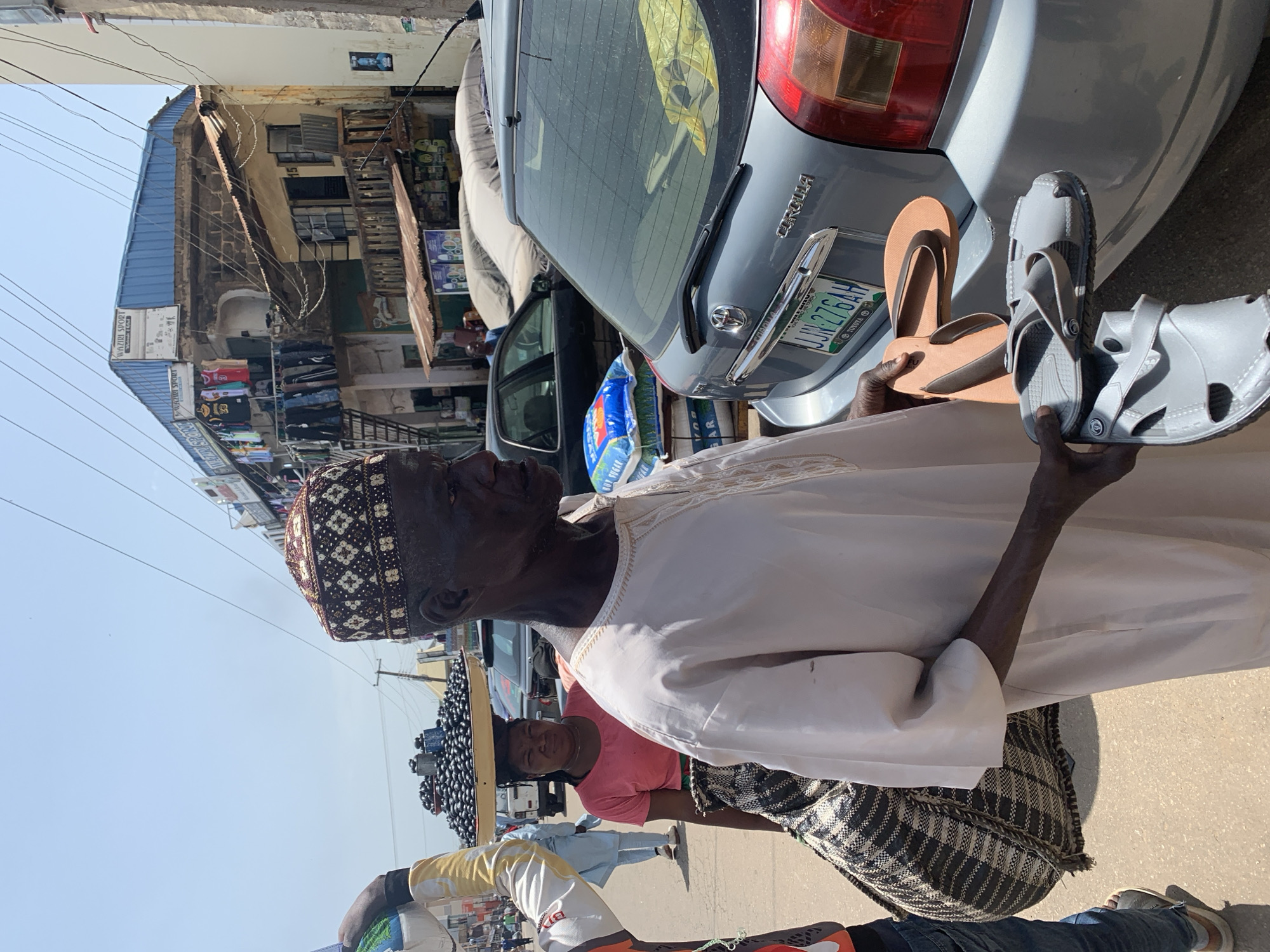The image depicts a street scene in Jos, Nigeria, with an elderly man in the foreground. He is wearing a traditional cap and a light-colored robe, and he is holding several pairs of sandals in his hands. Behind him, a woman is carrying a large tray of dark, round objects, likely food items, on her head. A gray Toyota Corolla is parked to the right of the frame, and in the background, there are multi-story buildings with various shops and hanging clothes, suggesting a commercial area. The sky is clear and bright, indicating a sunny day. The overall atmosphere appears to be that of a busy marketplace.