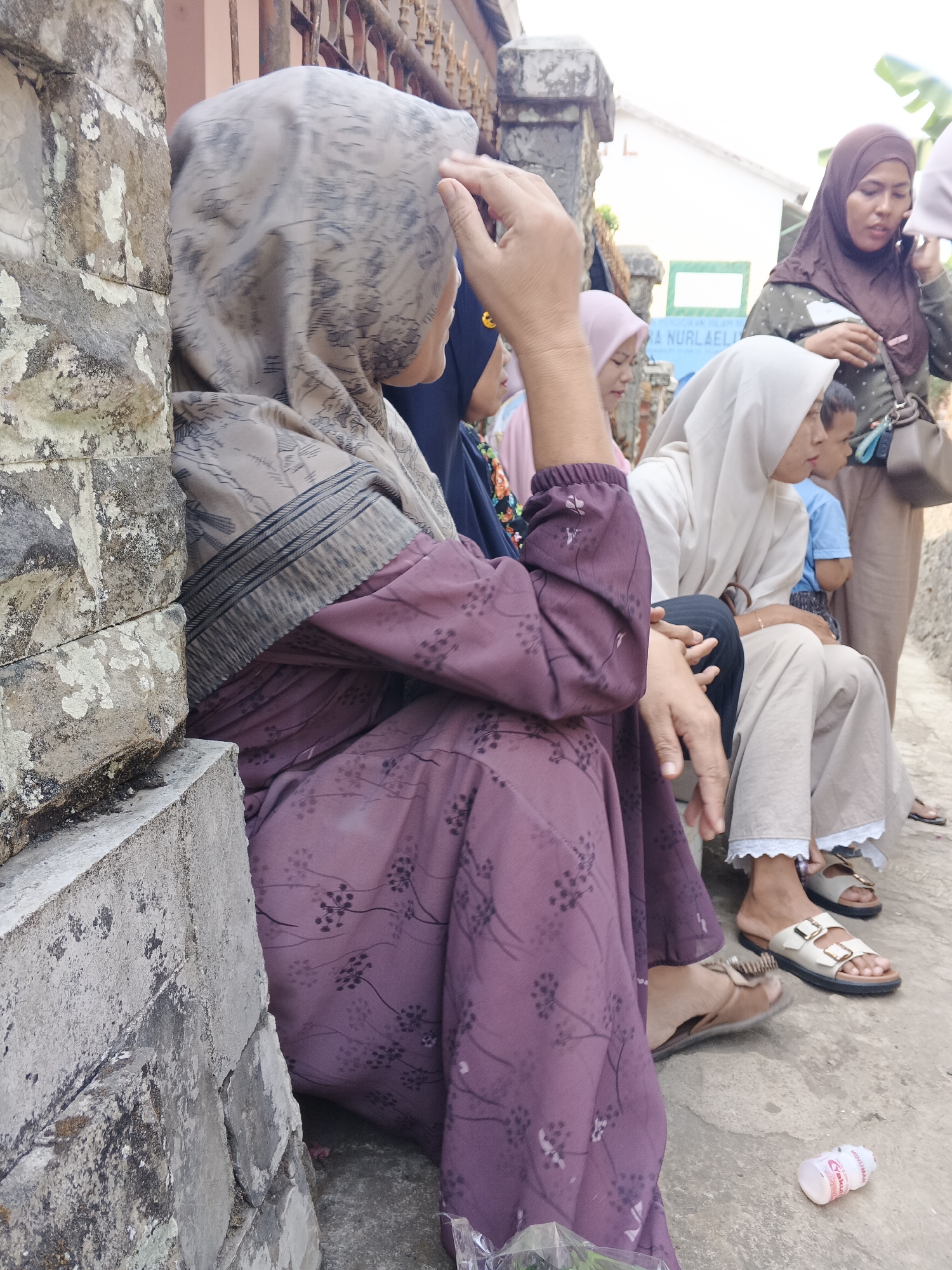 In Warungkiara, Indonesia, a group of people, primarily women wearing hijabs, are gathered outdoors near a stone wall and a weathered fence. The main subject, a woman in a purple patterned dress and a patterned hijab, is seated on a concrete ledge, her back mostly to the camera. She is touching her head with her right hand. To her right, a woman in a light beige hijab and pants is also seated. Further to the right, another woman in a dark brown hijab is standing and appears to be speaking on a mobile phone. Behind her, a child in a blue shirt is visible. The ground is paved with concrete, and a small, partially visible bottle of yogurt drink lies on the ground. The environment suggests a casual gathering in a residential or public outdoor space.