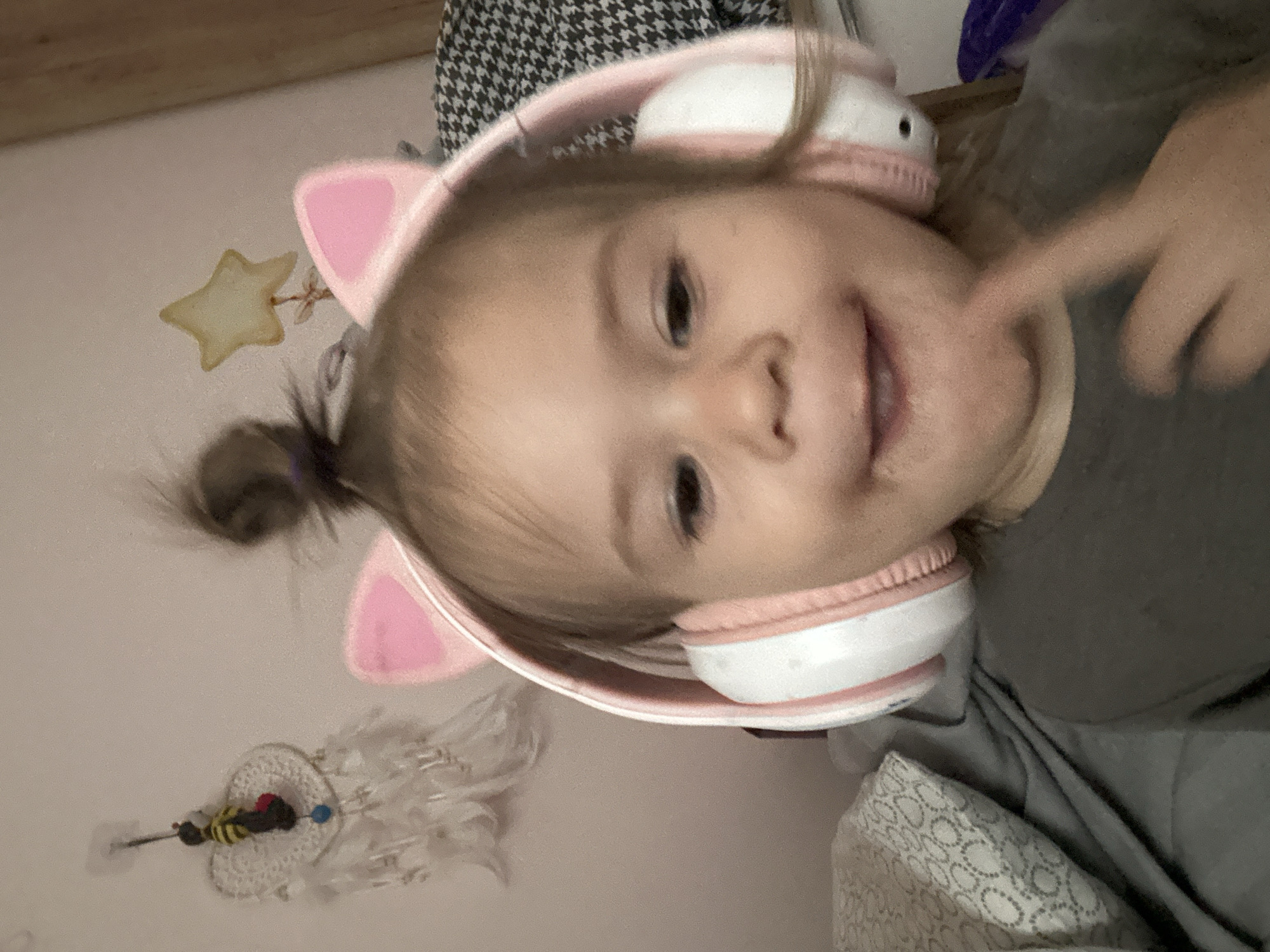 A child wearing headphones with cat ears is in an indoor setting. The child has brown hair, part of which is pulled back in a small ponytail. The child is smiling. The headphones are pink and white. The background includes a wall with decorations, including a star and a dreamcatcher. The child appears to be touching their cheek with a finger. The location is Ольховка, Russia.