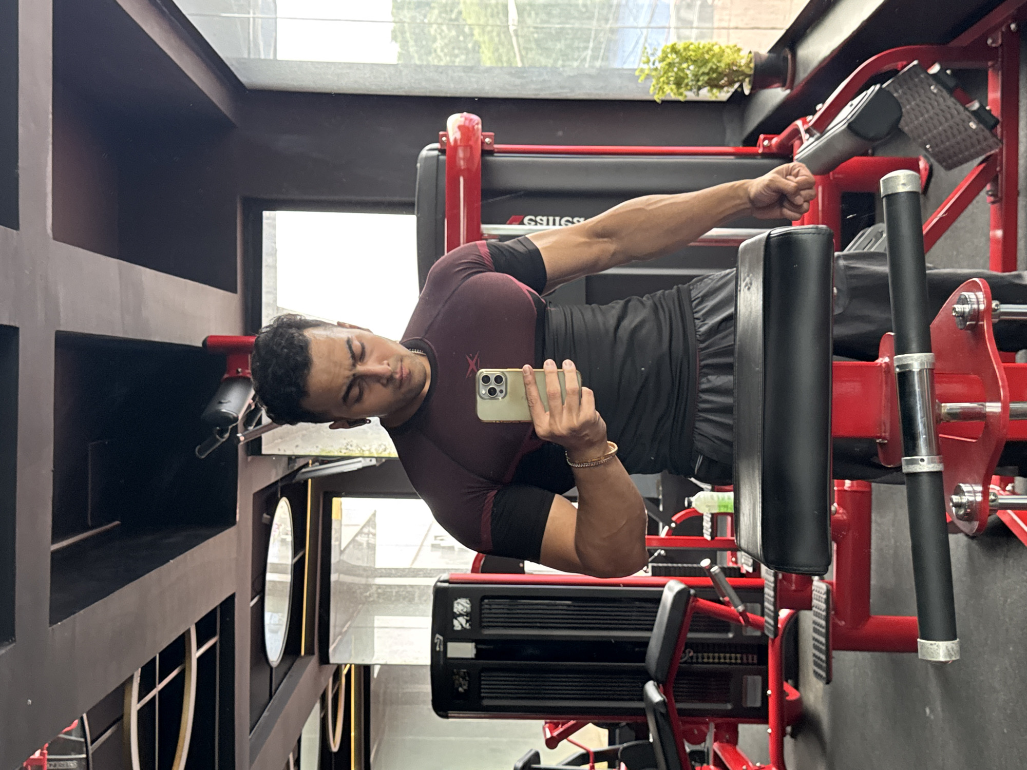 A man is taking a selfie in a gym, reflected in a phone. He is wearing a dark red and black t-shirt and dark shorts. He is standing in front of gym equipment, with a window to his right showing greenery outside. The gym has a modern industrial design with red and black accents. The scene appears to be located in Kolkata, India, as indicated by the specific gym equipment and the architectural style.