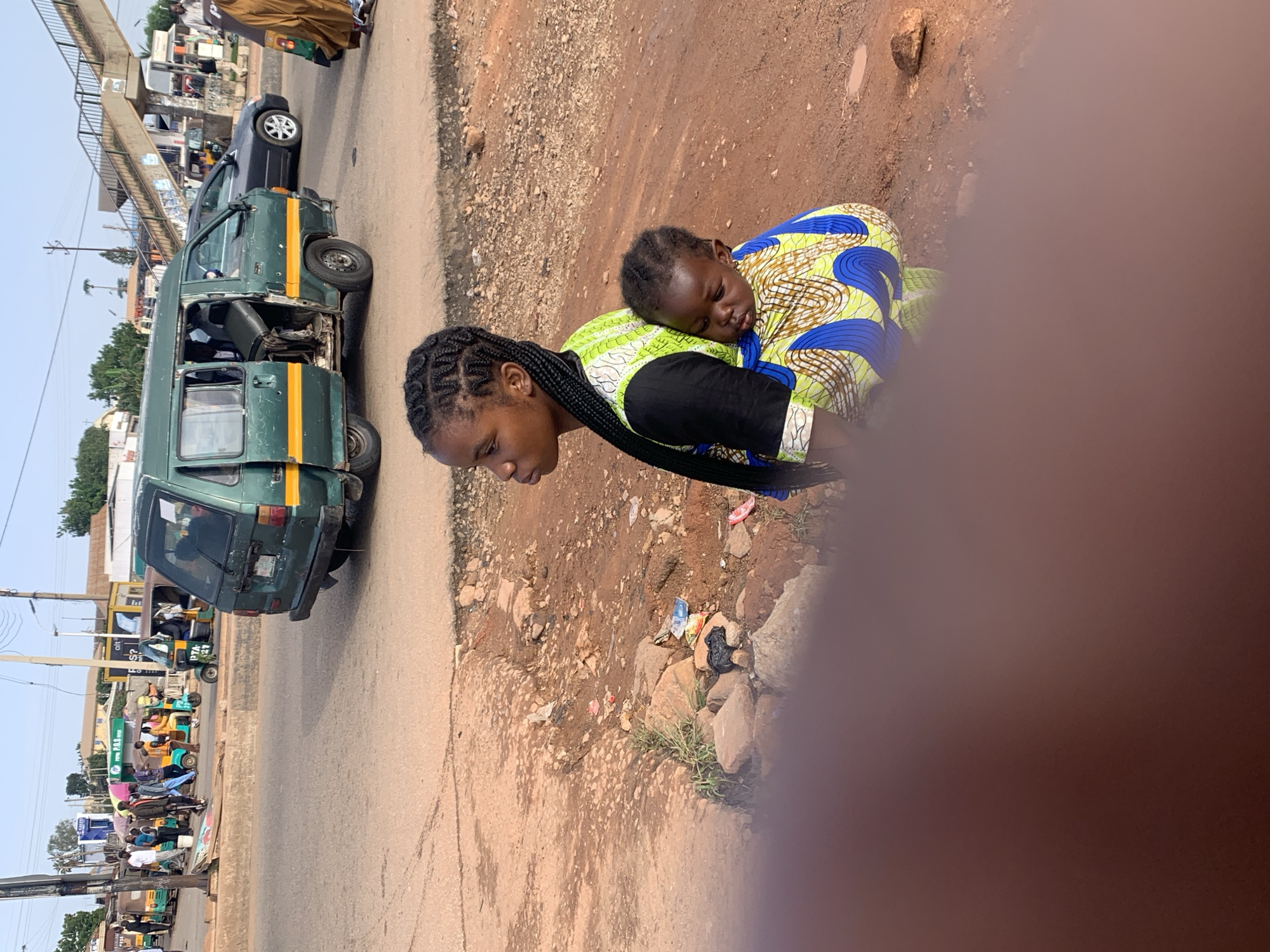The image depicts a street scene in Gura Top, Nigeria. In the foreground, a young woman is seated, carrying a baby on her back wrapped in a colorful cloth with a blue and yellow swirl pattern. The woman has her hair braided in a style of cornrows. She is wearing a black top with a lime green and white patterned vest over it. The ground around them is dirt and rocks, with some debris scattered about.
In the background, a green van with its door open is parked on a paved road. The van has yellow stripes along its side. Other vehicles, including several auto-rickshaws, are visible on the road, some with passengers. Pedestrians are also present, walking on the sidewalk and near the vehicles. A pedestrian bridge crosses over the road in the distance. The overall environment appears to be a busy urban or semi-urban area.