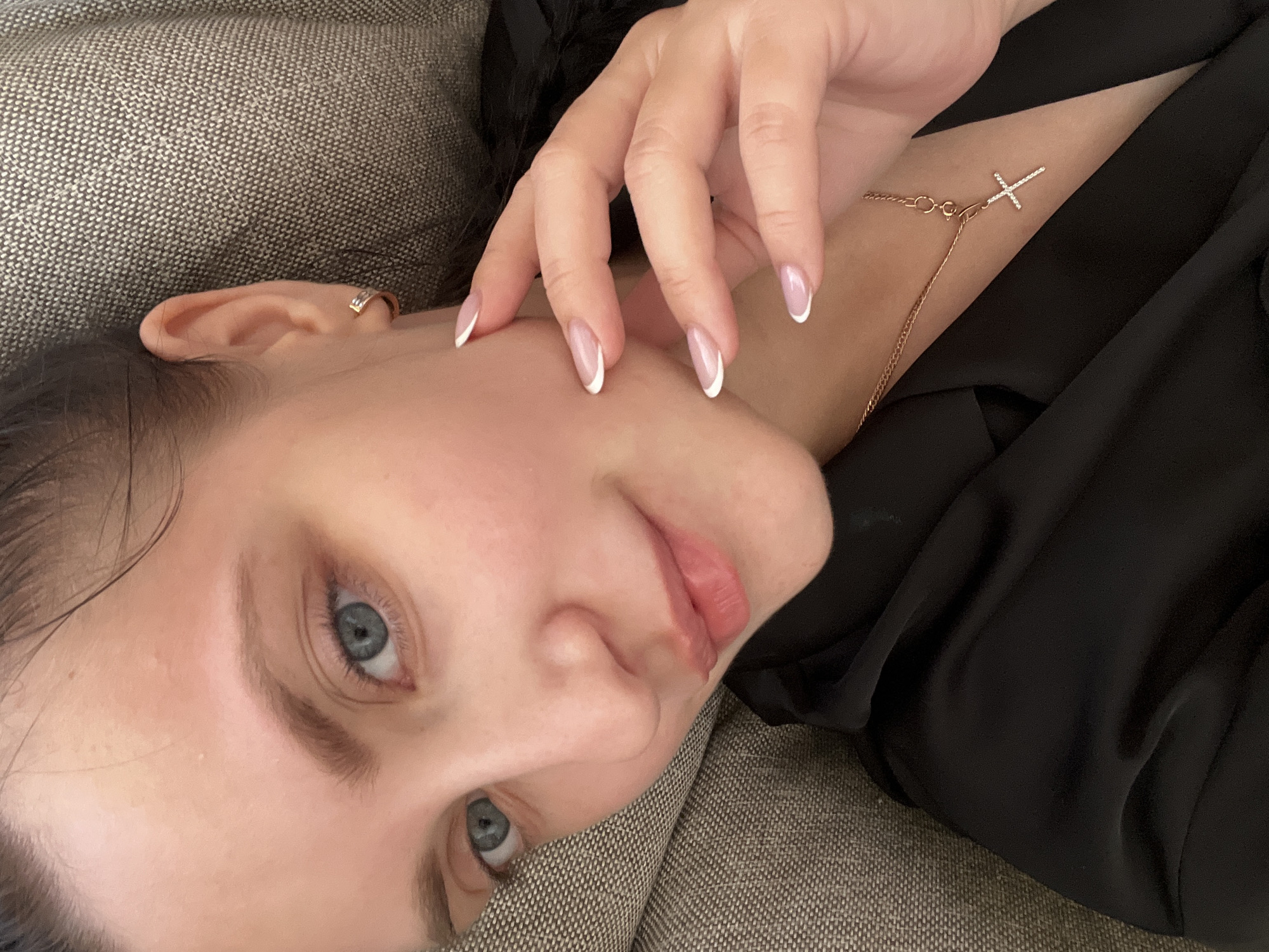 A close-up, slightly tilted portrait shows a young woman lying down in an indoor setting, likely on a sofa or bed, indicated by the textured, greyish-brown fabric visible in the background and foreground. She is looking directly towards the viewer with a calm and relaxed expression.
The woman has light skin, blue-grey eyes, and her dark hair is partially visible around her face and behind her ear. Her lips are slightly parted, revealing a soft pink color. She appears to be wearing minimal or natural makeup.
Her right hand, adorned with a neat French manicure featuring white tips on oval-shaped nails, is gently resting on her cheek and neck. A delicate, gold-colored chain necklace with a small, cross-shaped pendant, embellished with tiny white stones or diamonds, is visible on her neck. A small, gold-colored hoop or cuff earring is worn on the upper helix of her left ear. She is dressed in a black garment, possibly a top or robe, which has soft folds and drapes around her.
The overall mood of the image is tranquil and contemplative. No specific time of day or weather conditions can be discerned, but the lighting appears soft and natural, suggesting an indoor environment. There is no visible text in the image. The provided context indicates the location is Марковский, Russia, though there are no visual cues to confirm this within the frame.