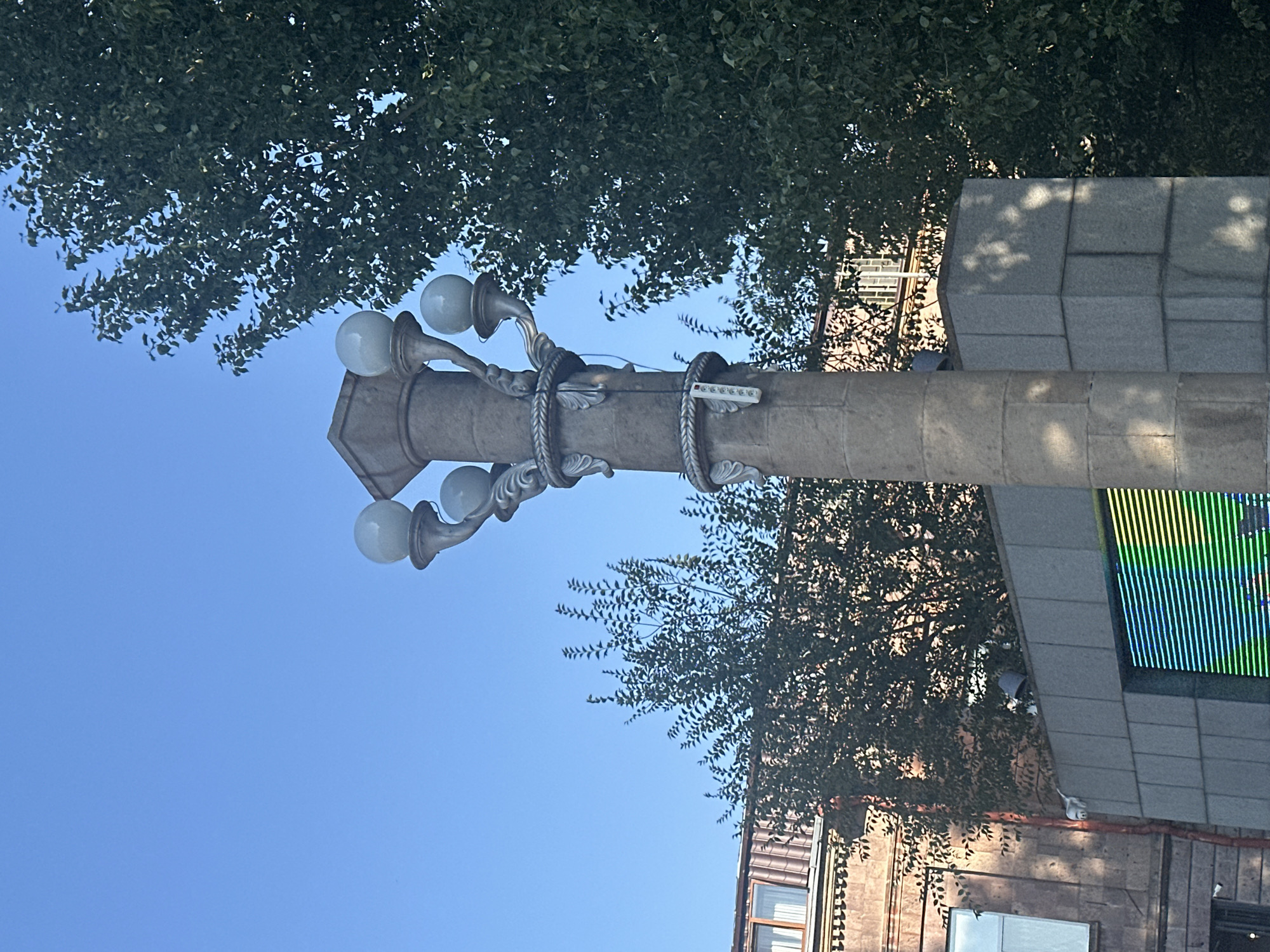 The image shows a street lamp with multiple globe lights, made of stone, in Gyumri, Armenia. The lamp is situated in an outdoor urban environment, with buildings and trees visible in the background. The sky is clear blue, indicating a sunny day. A digital display screen, showing horizontal green and yellow lines, is partially visible at the bottom of the frame. The scene appears to be a public space, possibly a square or a street. No people are visible in the image.