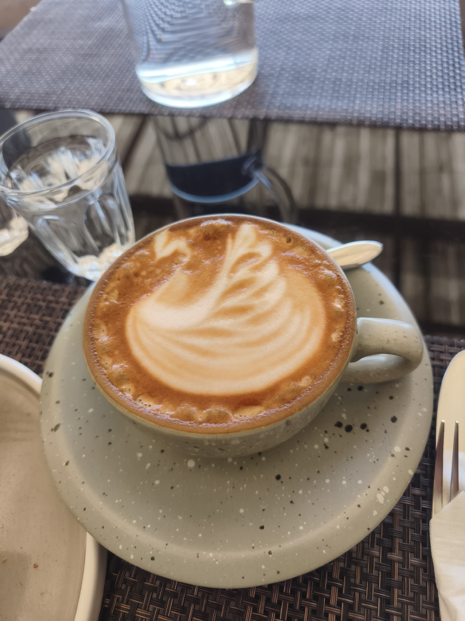 In Baan Mae Nam, Thailand, a close-up shot focuses on a coffee cup filled with latte art, placed on a speckled light grey saucer. The coffee features a creamy foam with a swirling pattern. A silver spoon rests on the saucer. To the left of the cup, a partially visible clear glass of water is present. In the background, another glass, also filled with water, sits on a woven placemat. The overall setting appears to be an outdoor dining area with a textured surface.