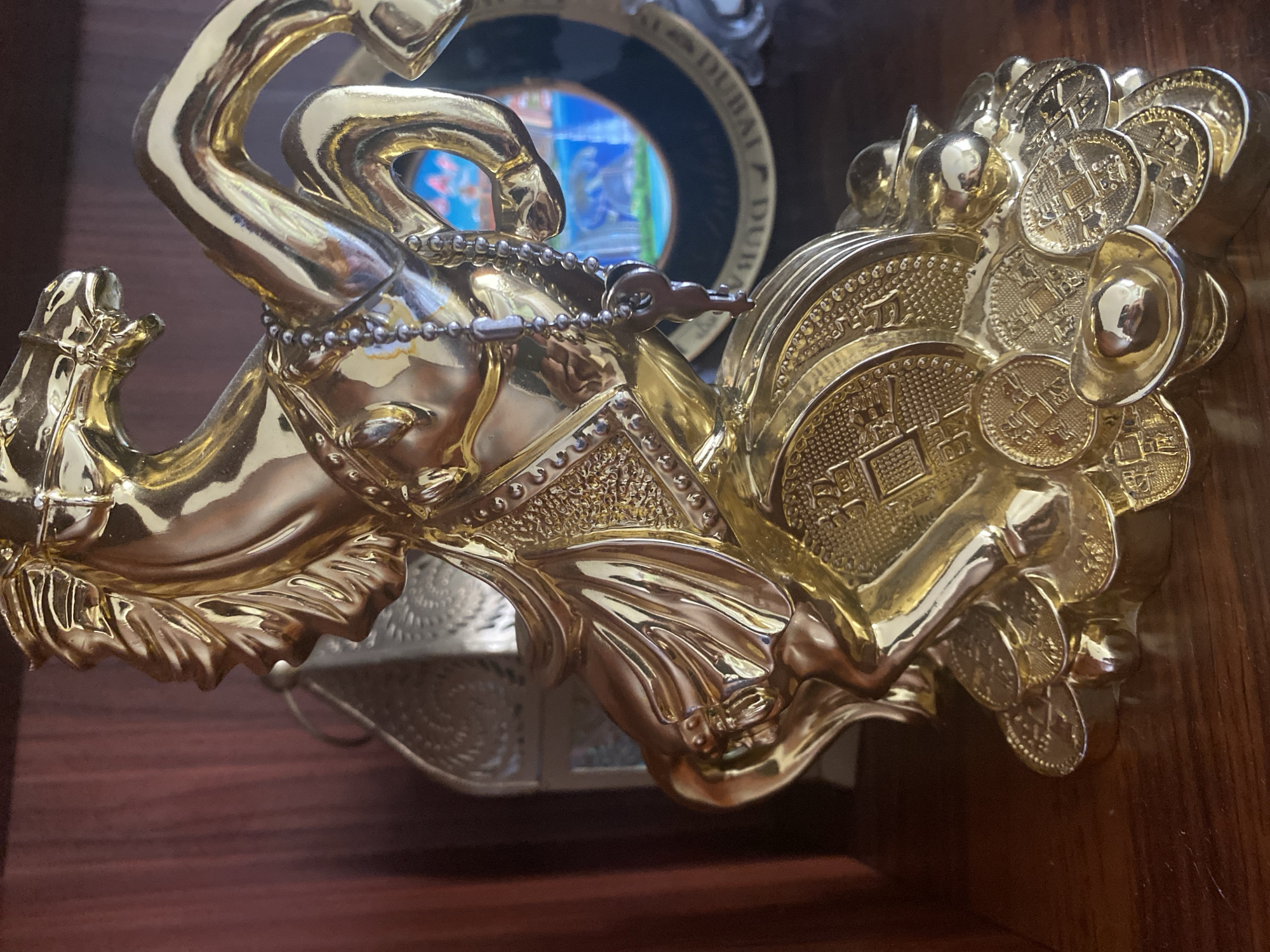 A close-up, eye-level shot shows a golden statue of a horse with intricate detailing, placed on a wooden surface. The horse is adorned with a beaded necklace and a small key charm. The base of the statue is composed of gold coins and ingots, some featuring Chinese characters. In the background, out of focus, is a silver lantern and a round, dark-colored decorative plate with a colorful image of a cityscape and water, possibly depicting Dubai based on visible text. The wooden surface appears to be a shelf.