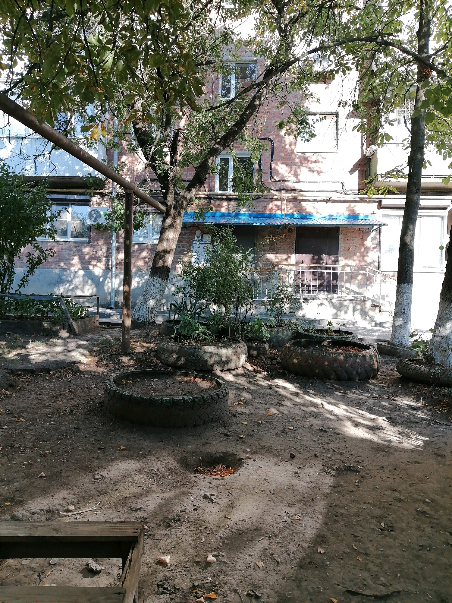 This image depicts a courtyard area with a brick building in the background. The building has multiple windows and a blue awning over an entrance. The courtyard is unpaved, with dirt and fallen leaves on the ground. Several old tires have been repurposed as planters, containing sparse vegetation. A metal railing runs along a small set of stairs leading to the building's entrance. Trees with green leaves are present, casting shadows on the ground. In the foreground, a wooden structure, possibly a bench or a raised planter, is visible. The scene appears to be a residential or communal outdoor space in a city. Based on the context provided, this location is identified as Rostov-on-Don, Russia. There are no people visible in the image.