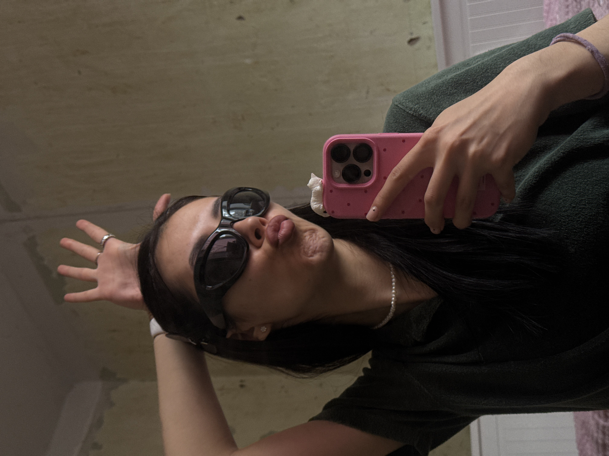 A woman is taking a selfie with a pink smartphone in what appears to be an indoor environment. She is wearing dark sunglasses and has her lips pursed in a kissing gesture. She has long dark hair and is wearing a pearl necklace and a bracelet. The background seems to be a wall with some disrepair or unfinished areas. There are no immediately apparent indications of the location being Votkinsk, Russia. The description is limited by the information visible in the image.