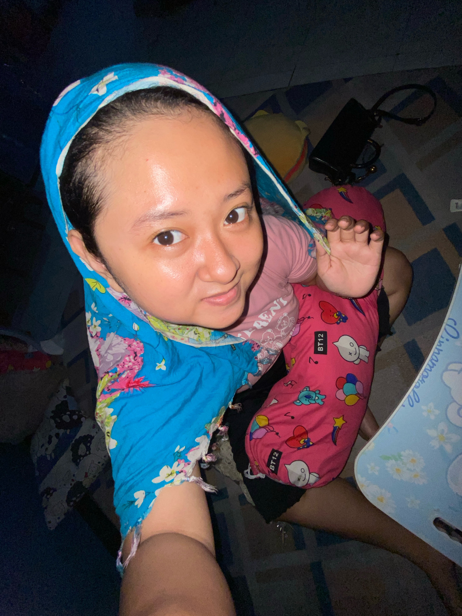 An overhead, high-angle, indoor photograph shows a person lying on the floor in Gelam, Indonesia. The subject, with fair skin and dark hair, wears a pink t-shirt and pink shorts decorated with cartoon characters and the letters "BT12." A blue headscarf with a floral pattern, adorned with white, pink, and yellow flowers, covers the person's head and drapes down the left side of the frame. Their right hand is raised towards the camera, fingers splayed. In the background, a black handbag with a strap rests on a yellow pillow, which is positioned on a blue and white patterned floor. A light blue laptop stand with a floral design is visible in the right foreground. The lighting is dim, suggesting the photograph was taken in low-light conditions.