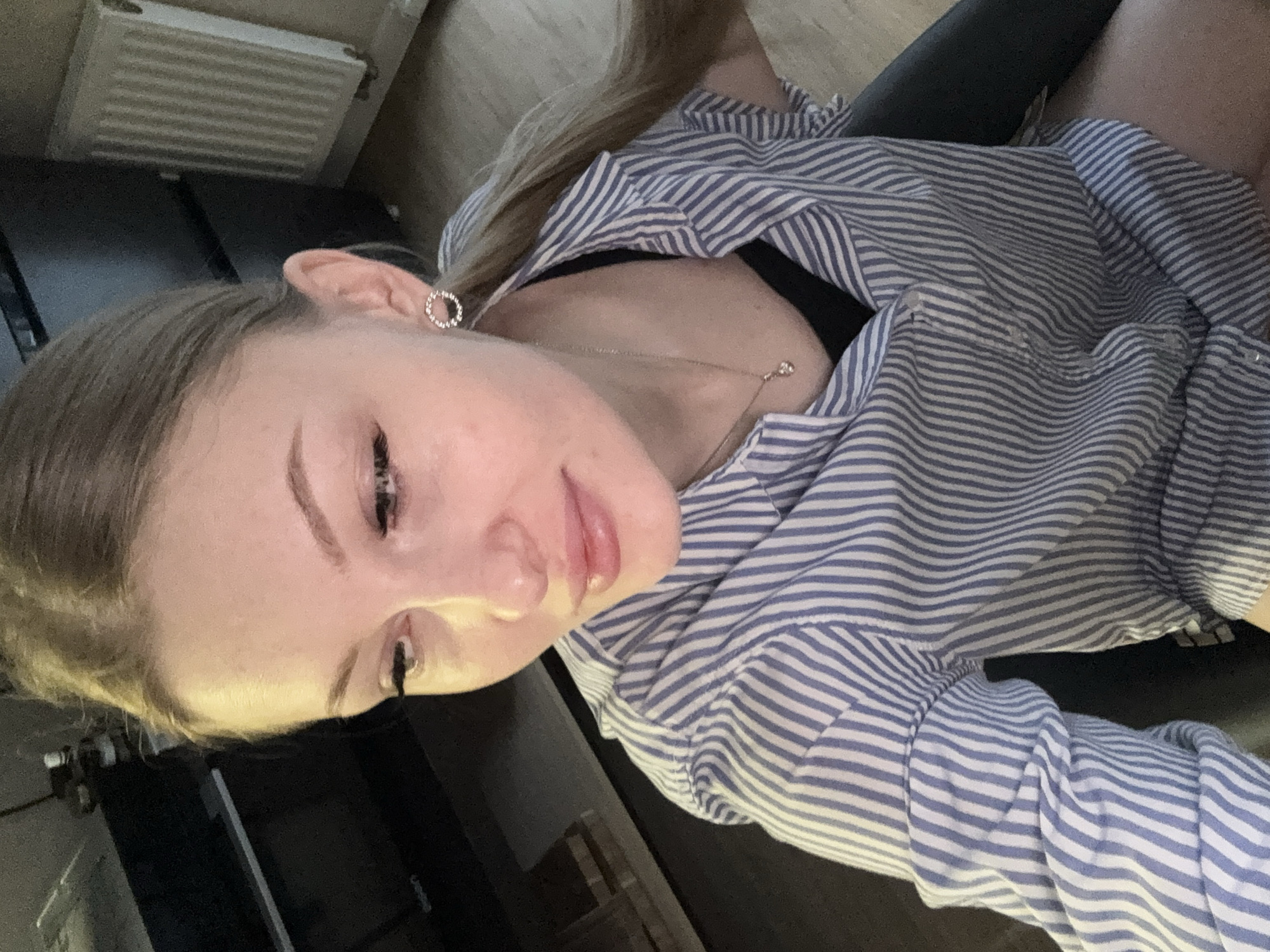 A young woman is seated indoors, angled slightly towards the camera. She has fair skin, light brown hair, and is wearing a blue and white striped shirt over a black garment. She has an earring and a delicate necklace is visible. The background includes a dark surface, possibly a desk or seat, and a radiator mounted on a wall. The image was taken inside an apartment, likely somewhere in Perm, Russia. There is no further information to specify the exact location.
