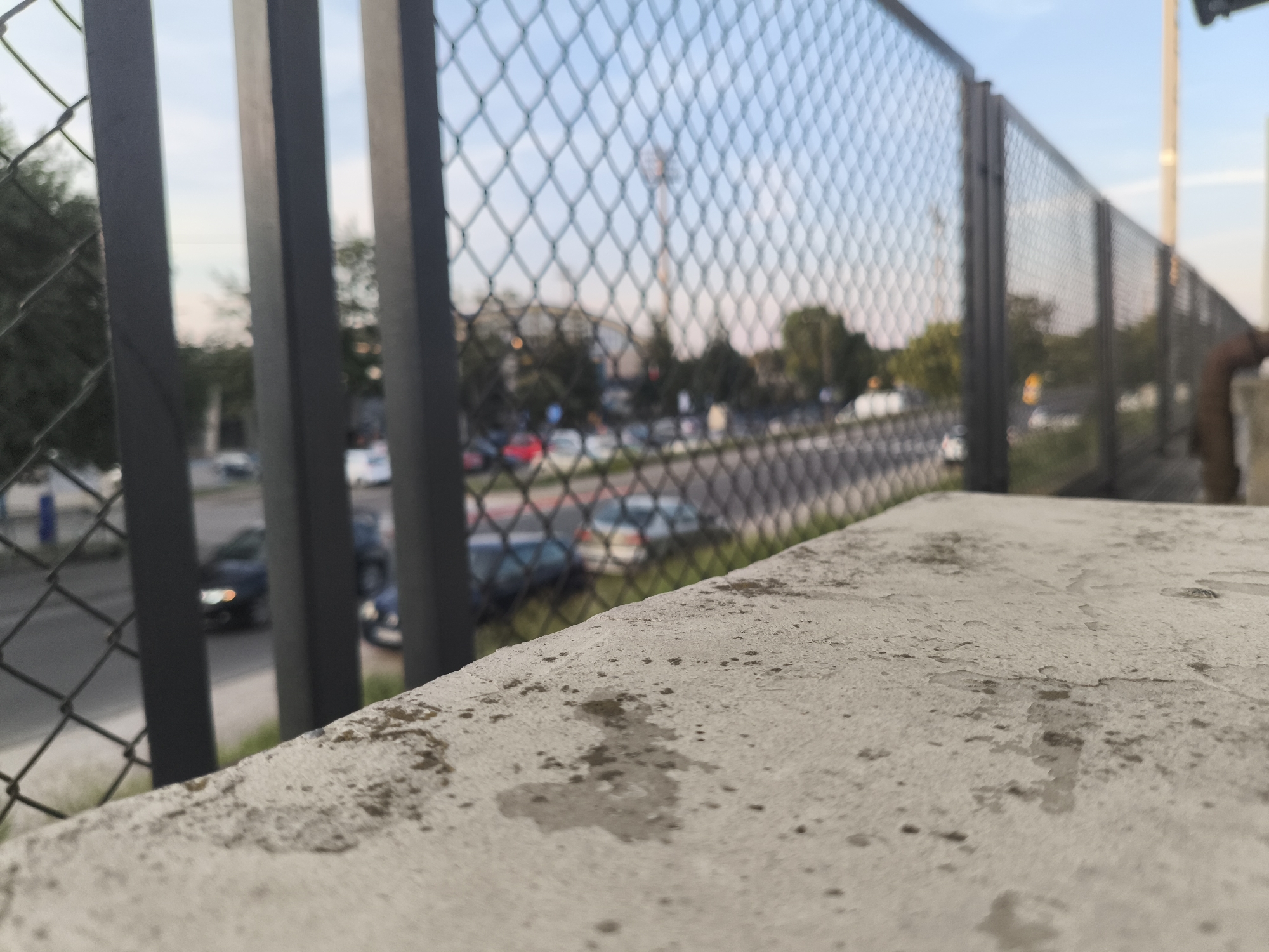 The image shows a close-up view of a rough, textured concrete surface in the foreground. Beyond this surface, a chain-link fence separates the viewer from a street scene with cars parked and driving. In the background, trees and buildings are visible, and a tall tower with lights can be seen in the distance. The sky above is a pale blue with some wispy clouds, suggesting it is either early morning or late afternoon. The overall impression is of an outdoor urban or suburban environment. The fence appears to be a safety barrier, possibly along a road or elevated walkway. The concrete surface seems to be part of a ledge or platform. No people are visible in the frame. The lighting suggests a soft, diffused light, consistent with either dawn or dusk. There is no visible text in the image.