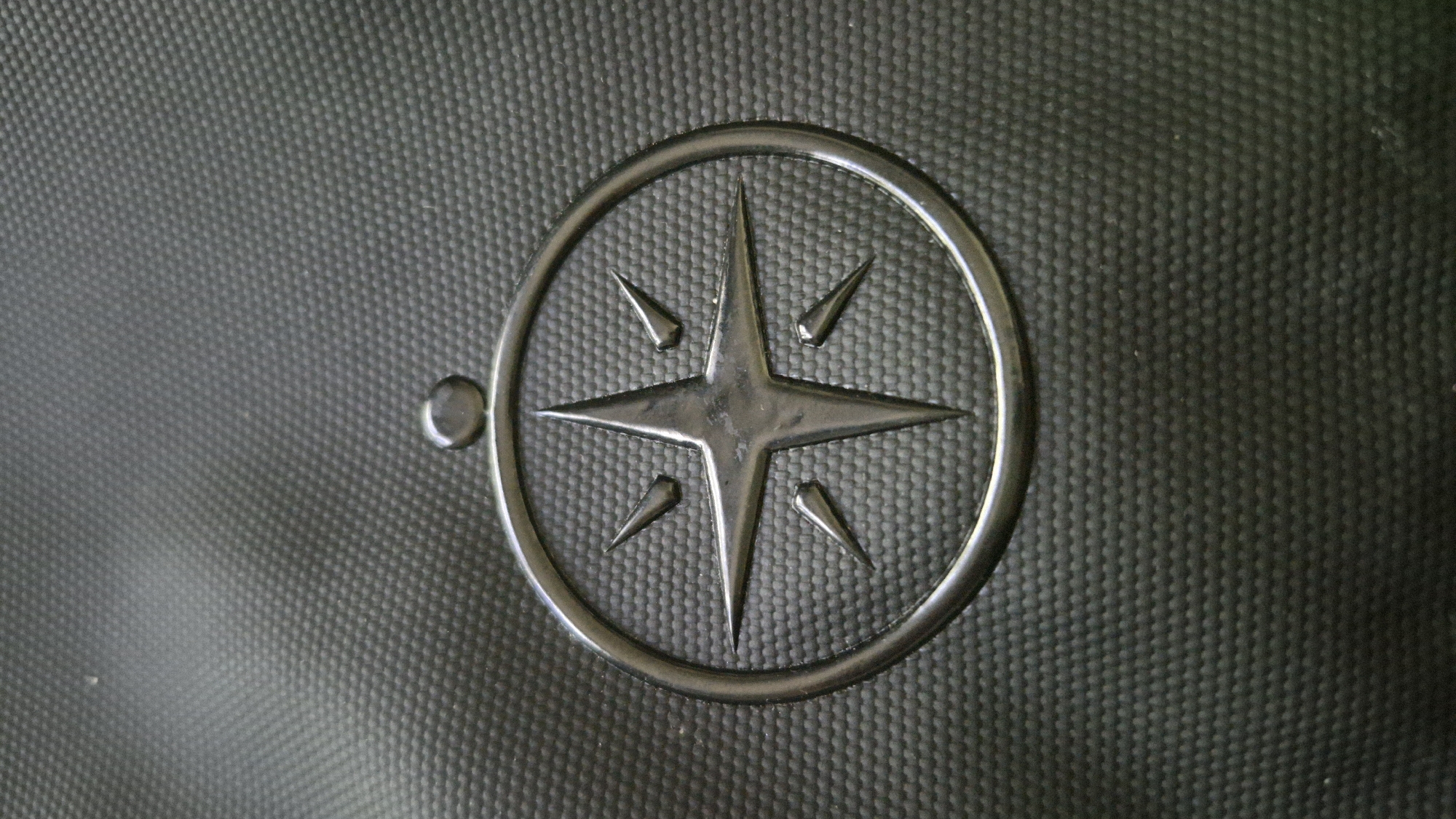 The image displays a close-up, high-angle shot of a dark, textured surface, possibly a fabric or synthetic material, with a raised, embossed compass rose logo in the center. The compass rose has eight points, with four longer, pointed tips alternating with four shorter, triangular points. A small, circular raised detail is positioned directly above the compass rose. The background consists of a uniform, pebbled texture. The lighting creates subtle highlights and shadows on the embossed elements, emphasizing their three-dimensional quality. There is no discernible location in the city of Gdańsk, Poland, evident in this image; it is a close-up of an object with a logo.