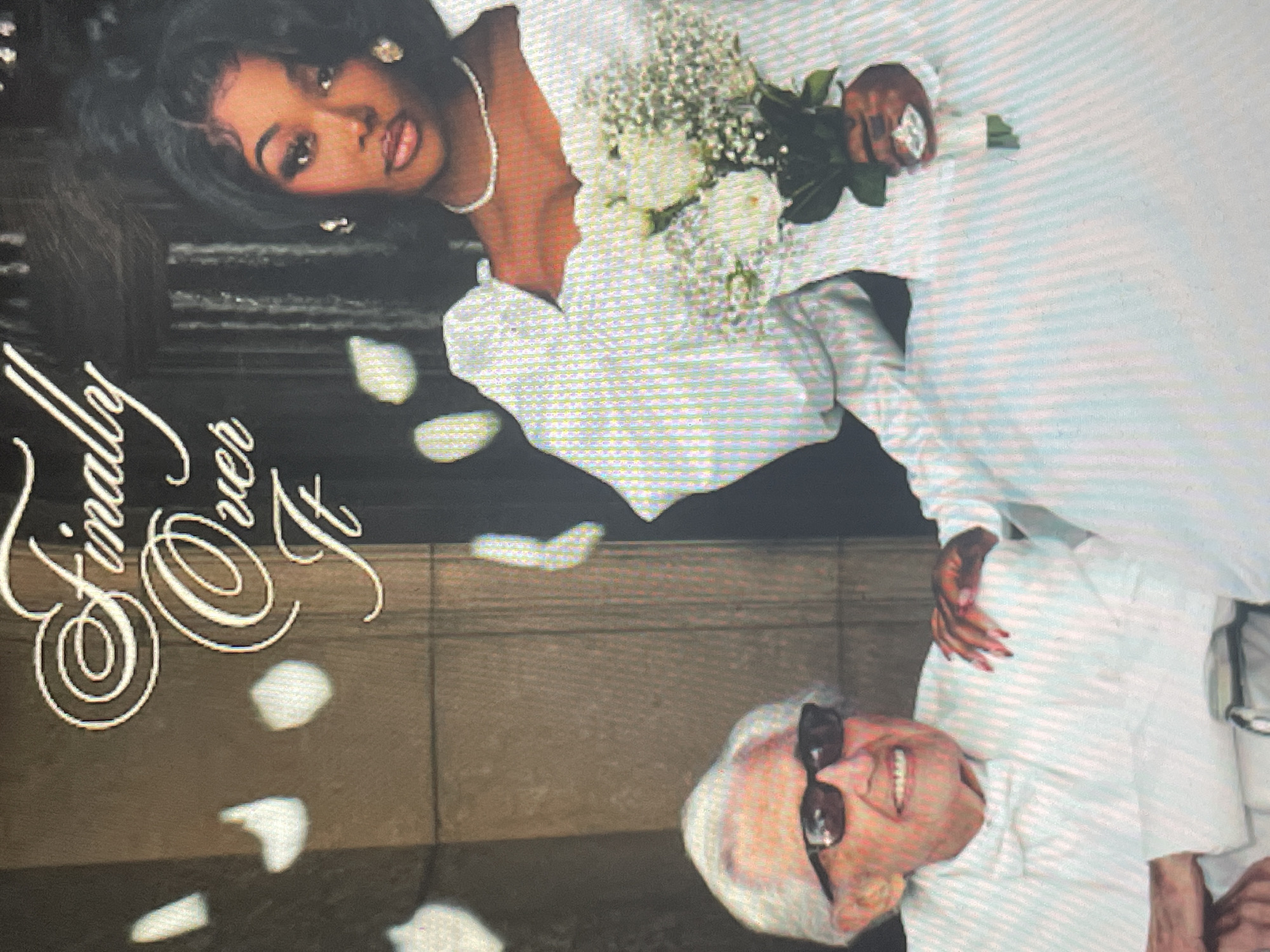 The image depicts a celebratory event, likely a wedding photoshoot, set against a dark background with scattered white flower petals. A dark-skinned woman with dark, curly hair, wearing a white long-sleeved wedding dress with ruffles, a pearl necklace, and pearl earrings, is positioned on the left. She holds a white bouquet of roses and baby's breath, and her fingernails are painted red. She looks directly at the viewer with a confident and composed expression. To her right, partially obscured, is an older, fair-skinned man with white hair, wearing a white shirt and dark sunglasses. He is smiling broadly, conveying joy. His hand, with red-painted nails, rests near his shoulder. The background is dark, featuring subtle horizontal lines and textures, possibly suggesting stone or concrete architecture. Prominently displayed in elegant, light-colored cursive script along the left edge of the image is the text "Finally Over It". The overall atmosphere suggests a significant life event or perhaps a creative visual for an album or project, given the prominent text. The image exhibits a slightly grainy or pixelated quality.