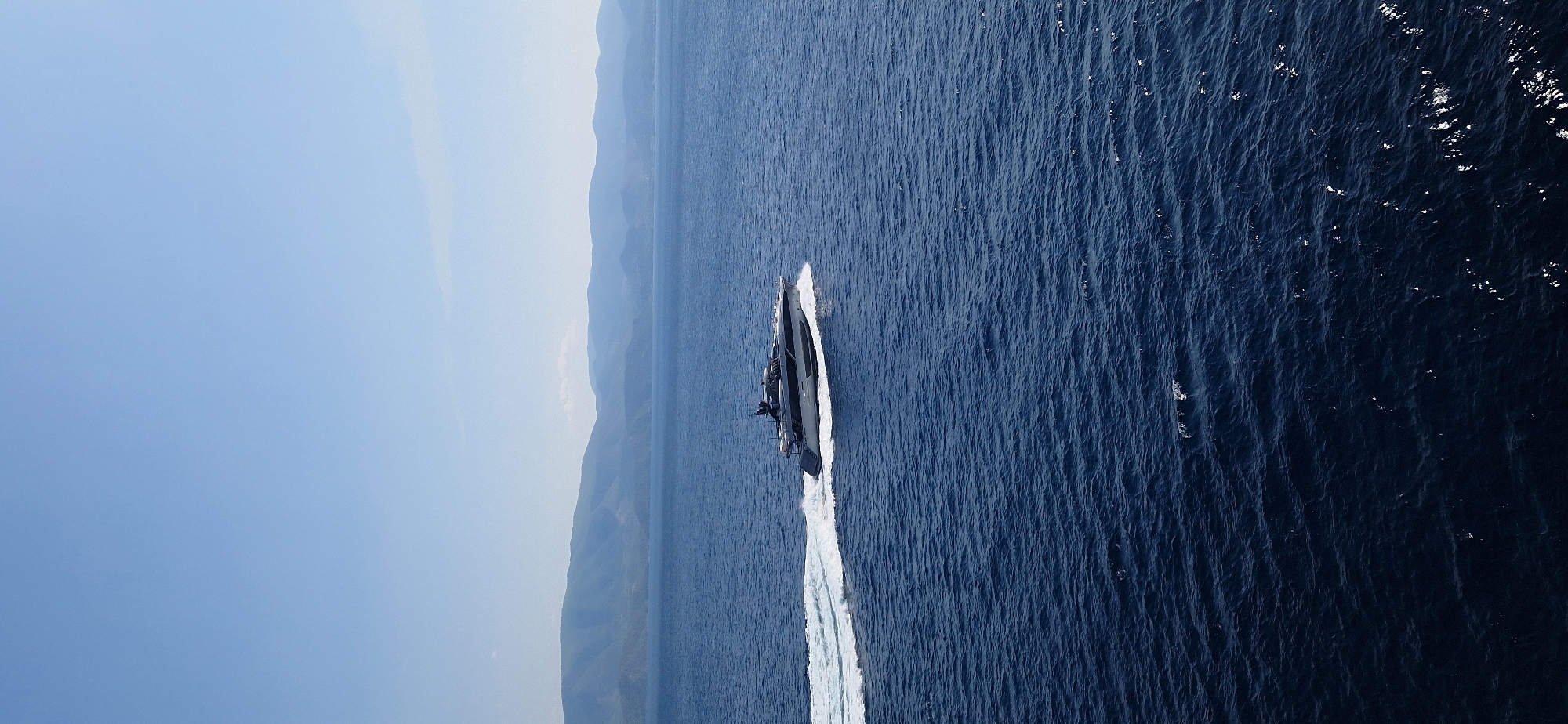 A modern, sleek silver and black yacht speeds through deep blue water, leaving a white wake behind it. The yacht is angled from the left to the right, with its bow facing towards the right of the frame. In the background, a line of hazy, blue-green mountains stretches across the horizon under a clear, light blue sky. The image is likely taken from an aerial perspective, looking down on the water and the distant coastline. The location is within the Municipal Unit of Agioi Theodoroi, Greece.