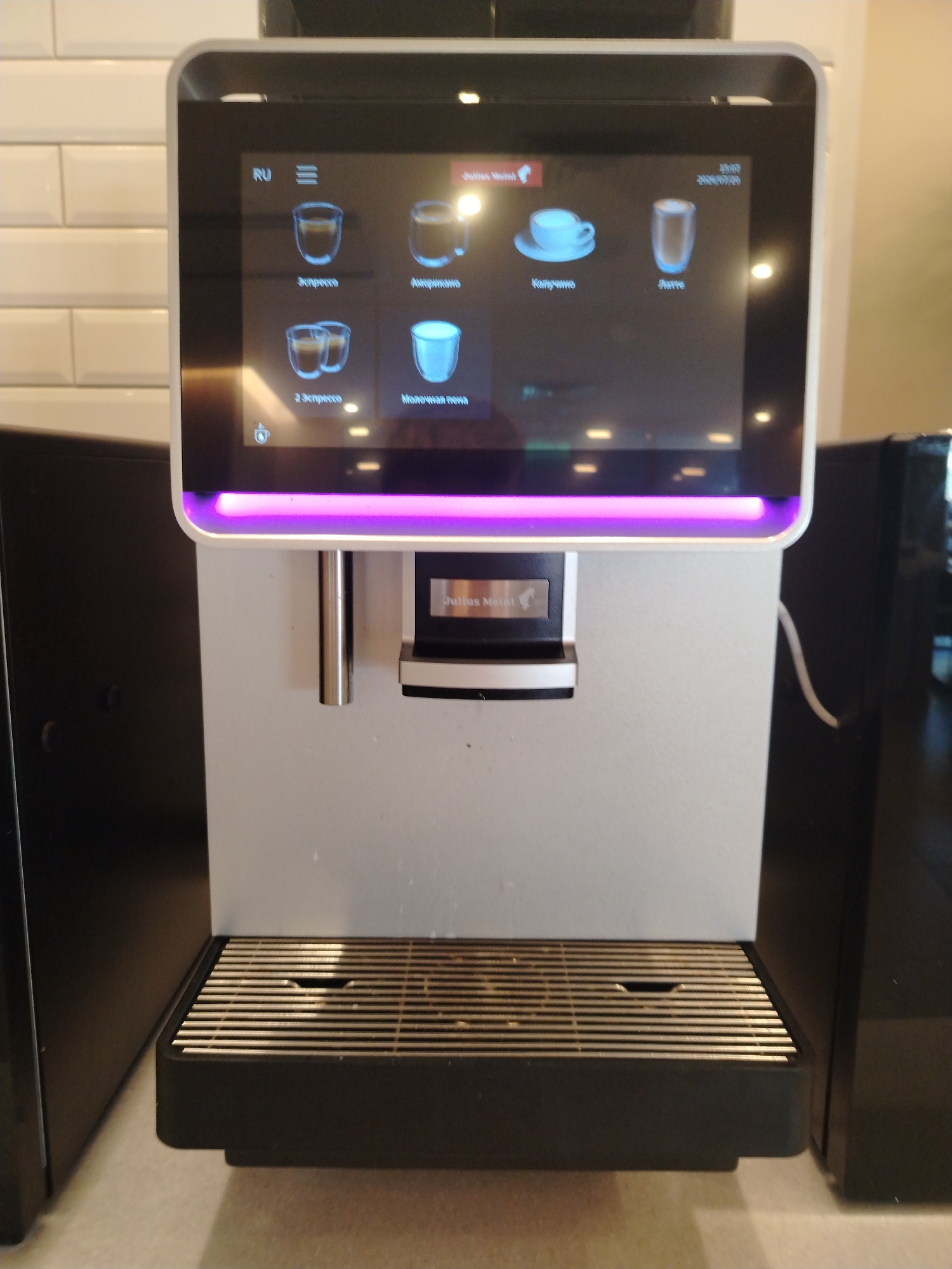 The image shows a modern, silver and black coffee machine with a touchscreen display. The touchscreen displays various coffee drink options with icons, including "Эспрессо" (Espresso), "Американо" (Americano), "Капучино" (Cappuccino), "Латте" (Latte), "2 эспрессо" (2 Espresso), and "Молочная пена" (Milk Foam). The machine has a silver drip tray with a metal grate. A white cable is visible plugged into the back of the machine. The background consists of a white tiled wall on the left and a black surface on the right, suggesting it is in a commercial or domestic kitchen setting. The coffee machine is the primary subject, and it appears to be in a resting state, with the display illuminated.