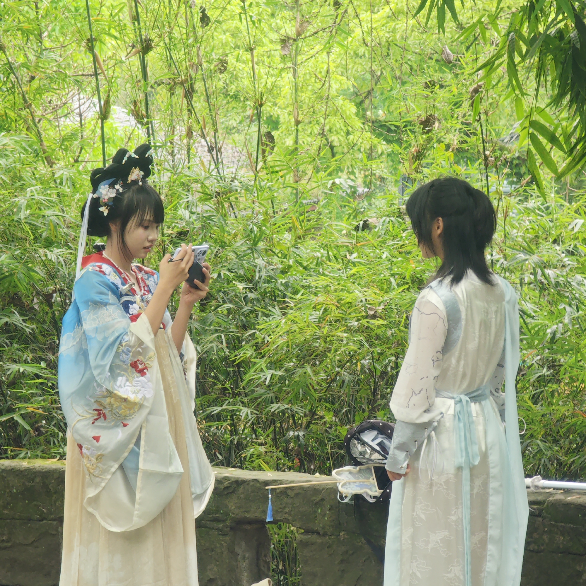 In Yuzhong District, China, two young women stand in front of a dense bamboo forest. The woman on the left is actively taking a picture with her phone, while the other woman is positioned to be photographed. Both women are dressed in traditional Chinese Hanfu clothing. A bridge is visible in the background. A bag and a sword are placed on the bridge.