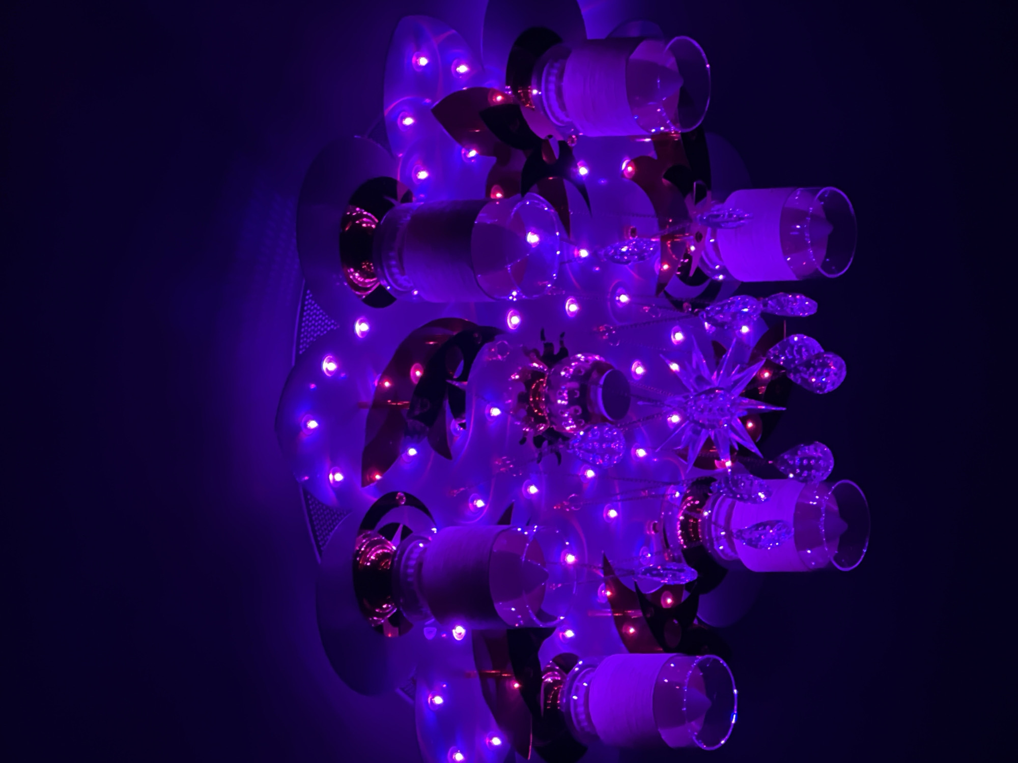 A close-up, tilted, medium-shot of a decorative lighting fixture against a dark, purple-toned background, location unknown. The fixture appears to be a complex assembly of various materials and shapes, illuminated with numerous small, bright, purplish-pink LED lights. The structure includes: reflective disks with cut-out geometric shapes; cylindrical clear glass or plastic cups attached horizontally; decorative metallic elements in copper or bronze tones shaped like stars; and hanging crystal-like decorations. The overall effect is a complex, visually stimulating display. The image appears to be shot indoors, possibly in a bar or entertainment setting.
