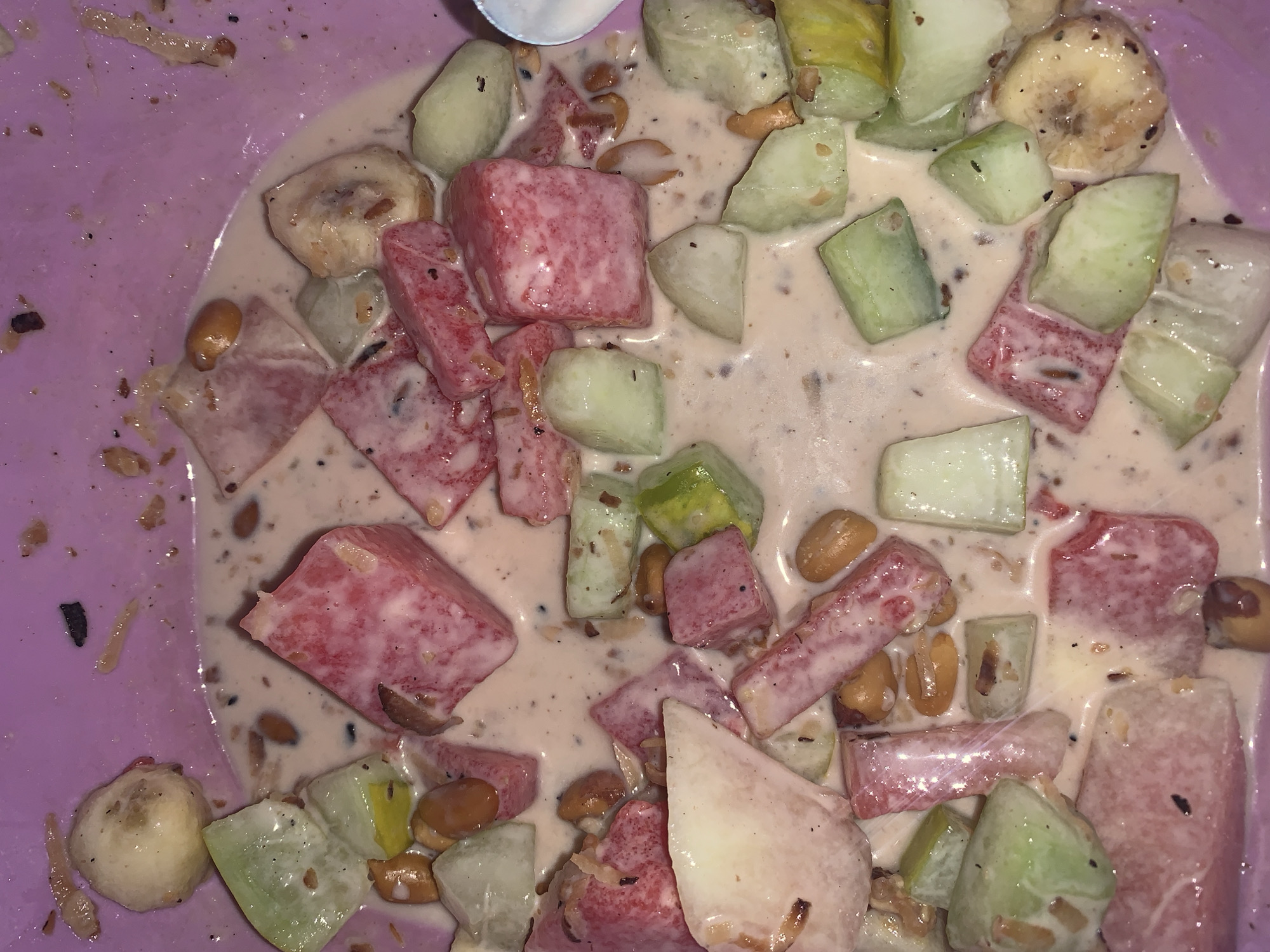 This close-up, top-down image displays a bowl filled with a colorful fruit and nut mixture submerged in a creamy, off-white liquid, set against a light purple container. The primary subjects are various chopped fruits, including prominent red and pink watermelon cubes, light green cucumber pieces, and several round slices of banana, some of which exhibit browning. Small, light brown, oval-shaped nuts, likely groundnuts or peanuts, are scattered throughout the mixture.
The creamy liquid appears thick and is speckled with numerous small dark particles, possibly spices or ground nuts, along with some lighter, elongated fibrous remnants. A small section of a white spoon is visible near the top edge of the frame. The scene captures a prepared food item, resembling a fruit salad or a dessert common in certain regions. The fruits appear slightly softened by the creamy dressing. No text is visible. The location context for this dish is Rafin Par, Nigeria.