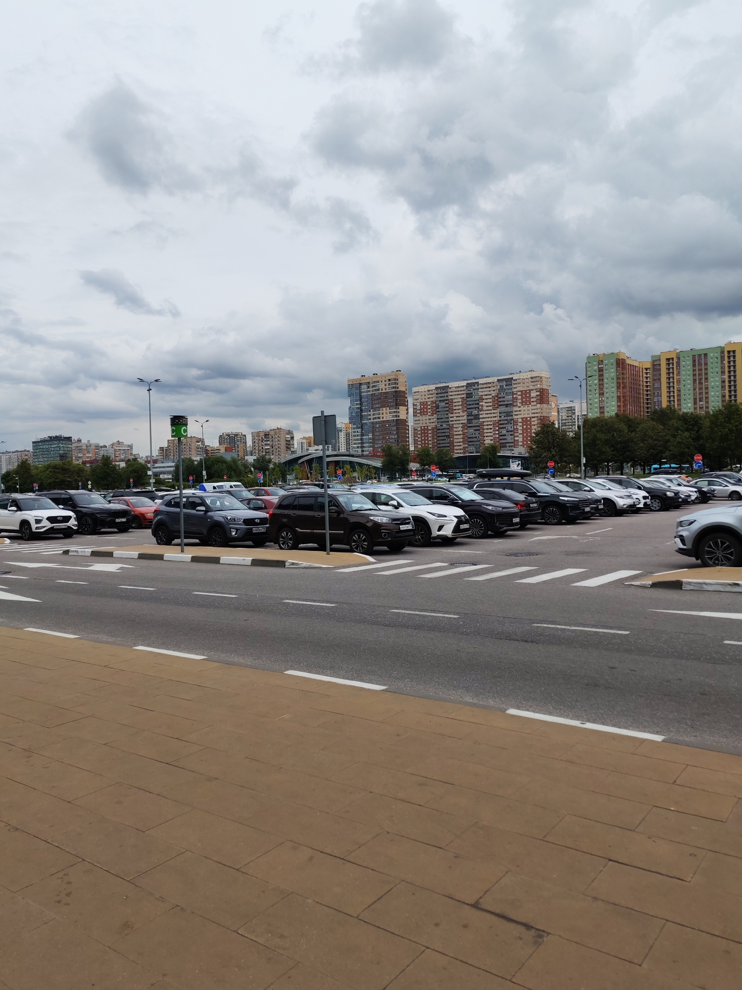 A cloudy day is depicted in this outdoor photograph, showcasing a row of parked cars in what appears to be a parking lot. The cars are varied in color and model, including SUVs and sedans. In the background, several modern, multi-story buildings with colorful facades are visible, suggesting an urban or suburban setting. A paved walkway runs in the foreground, bordered by a road with white lane markings. A traffic signal with green and white lights is positioned near the cars. No people are visible in the image.