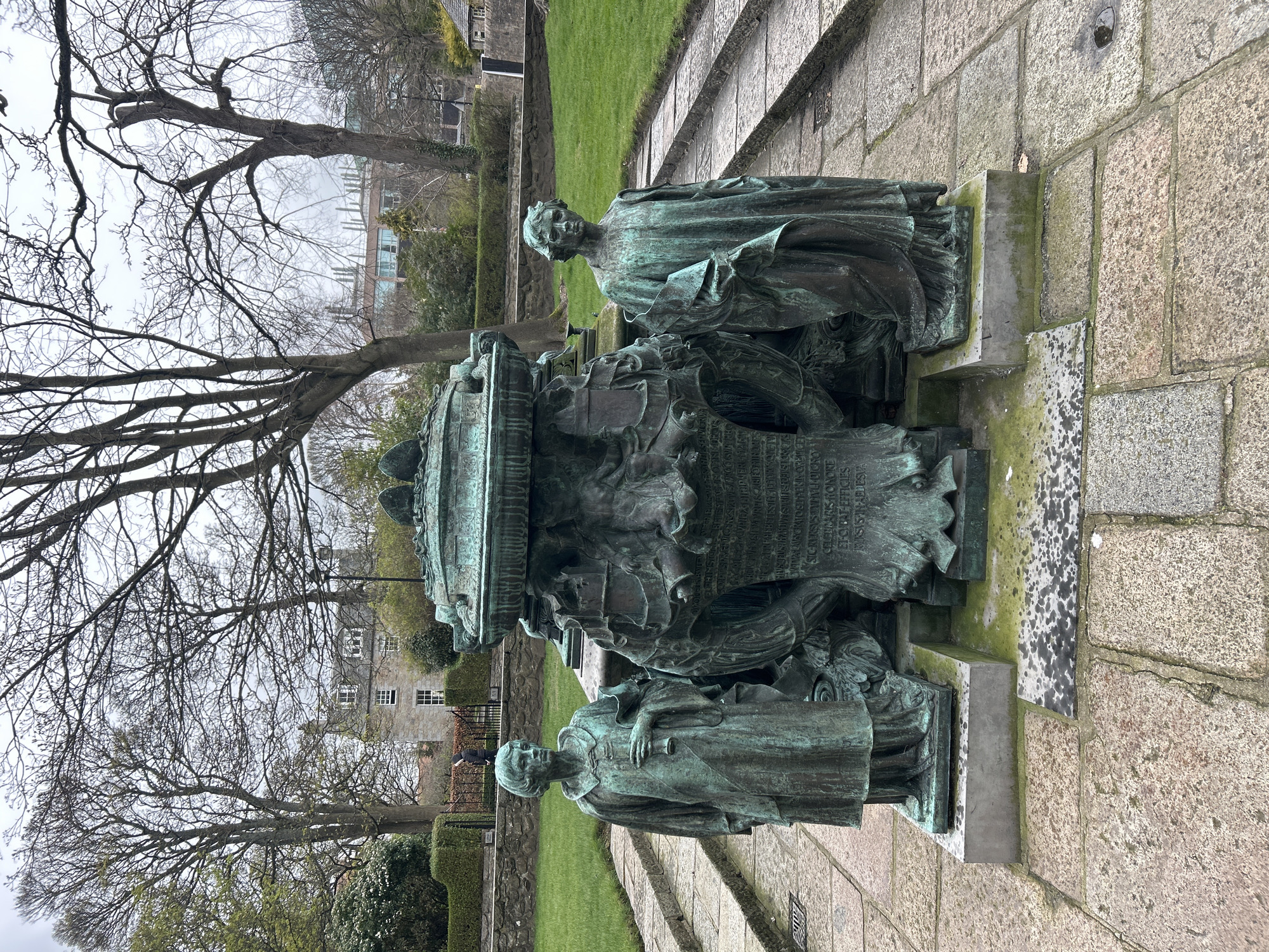Here's a description of the image:
The image shows the William Kelly Memorial in Aberdeen, United Kingdom. The monument is made of bronze and features two statues of women standing on either side of a large central structure covered in intricate carvings and text. The statues and structure are mounted on a stone base.
The monument sits on a paved area with grass and trees surrounding it. In the background, there are buildings and foliage. The sky appears overcast.