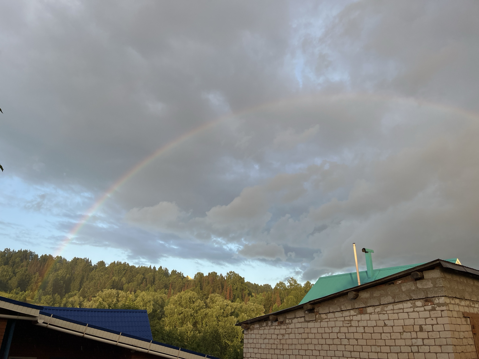 A rainbow arches across a cloudy sky over a rural landscape. In the foreground, the roofs of two buildings are visible. The building on the left has a dark roof, while the building on the right has a teal roof and is constructed of white brick. Beyond the buildings, a dense forest covers rolling hills. The image appears to have been taken in Olkhovka, Russia.