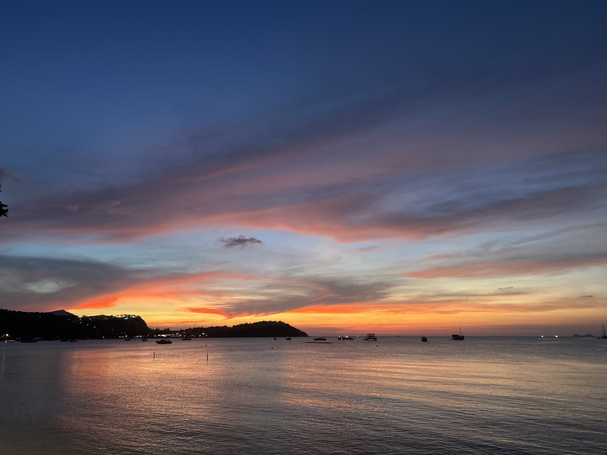 The image depicts a sunset over the water in Baan Bang Rak, Thailand. The sky is a mix of deep blue, light blue, pink, gray, and orange hues. Clouds stretch across the sky horizontally. The water reflects the colors of the sunset. The land is visible in the distance, silhouetted against the horizon. There are several boats on the water.