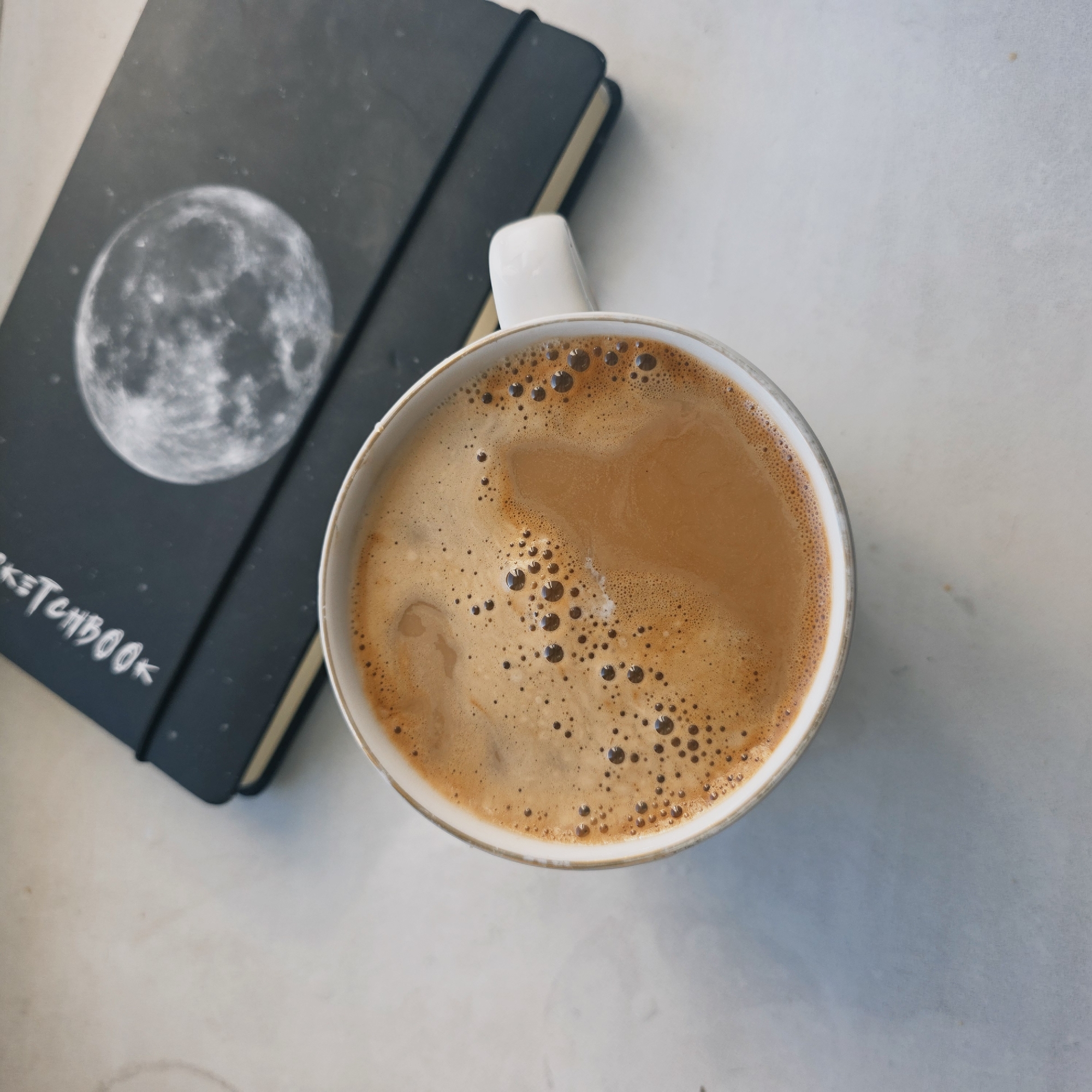 The image shows a cup of coffee next to a black sketchbook with a moon image on its cover. The sketchbook has the word "SKETCHBOOK" written on it in white. The coffee has a light brown color with darker brown bubbles on the surface. The background is a light, mottled surface. There is no evidence to suggest this image was taken in Saint Petersburg, Russia. There are no people visible in the image.