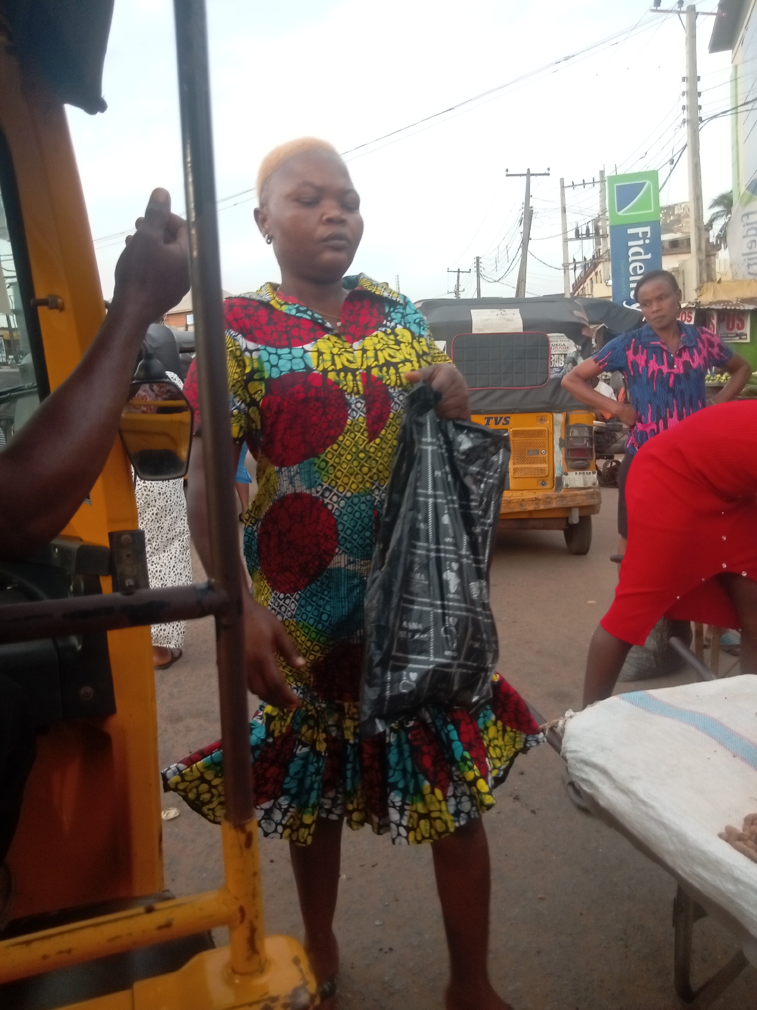 The image depicts a street scene in Nkpor, Nigeria. A woman with bleached blonde hair and wearing a colorful patterned dress stands holding a black plastic bag. She appears to be in front of a yellow auto rickshaw ("keke"). Partially visible within the auto rickshaw is a driver with dark skin. In the background, another woman in a blue shirt stands with her hands on her hips. To the right, a person in a red dress is bending over a table. A Fidelity Bank sign is also visible in the background, along with utility poles and wires.