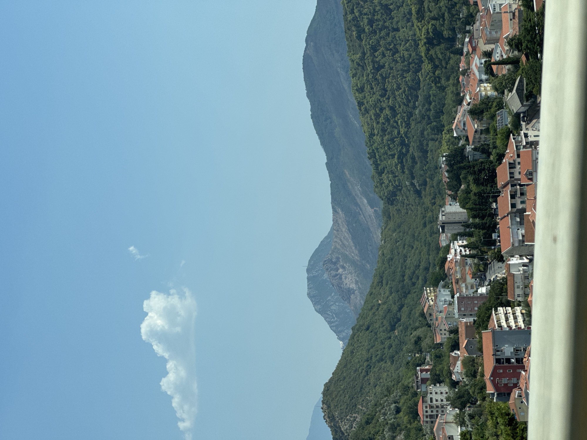 This is a photograph of the city of Nerin, Montenegro, taken from a high vantage point. In the foreground, a white railing is visible. The middle ground is filled with a dense collection of red-tiled rooftops and buildings, varying in height and architectural style, nestled on a verdant, tree-covered hillside. Beyond the city, a series of rolling green mountains dominate the landscape, with a larger, more rugged, and rocky mountain peak in the distance. The sky is a clear, pale blue with a single, small white cloud in the upper left quadrant. The overall scene depicts a coastal town situated within a mountainous terrain on a sunny day.