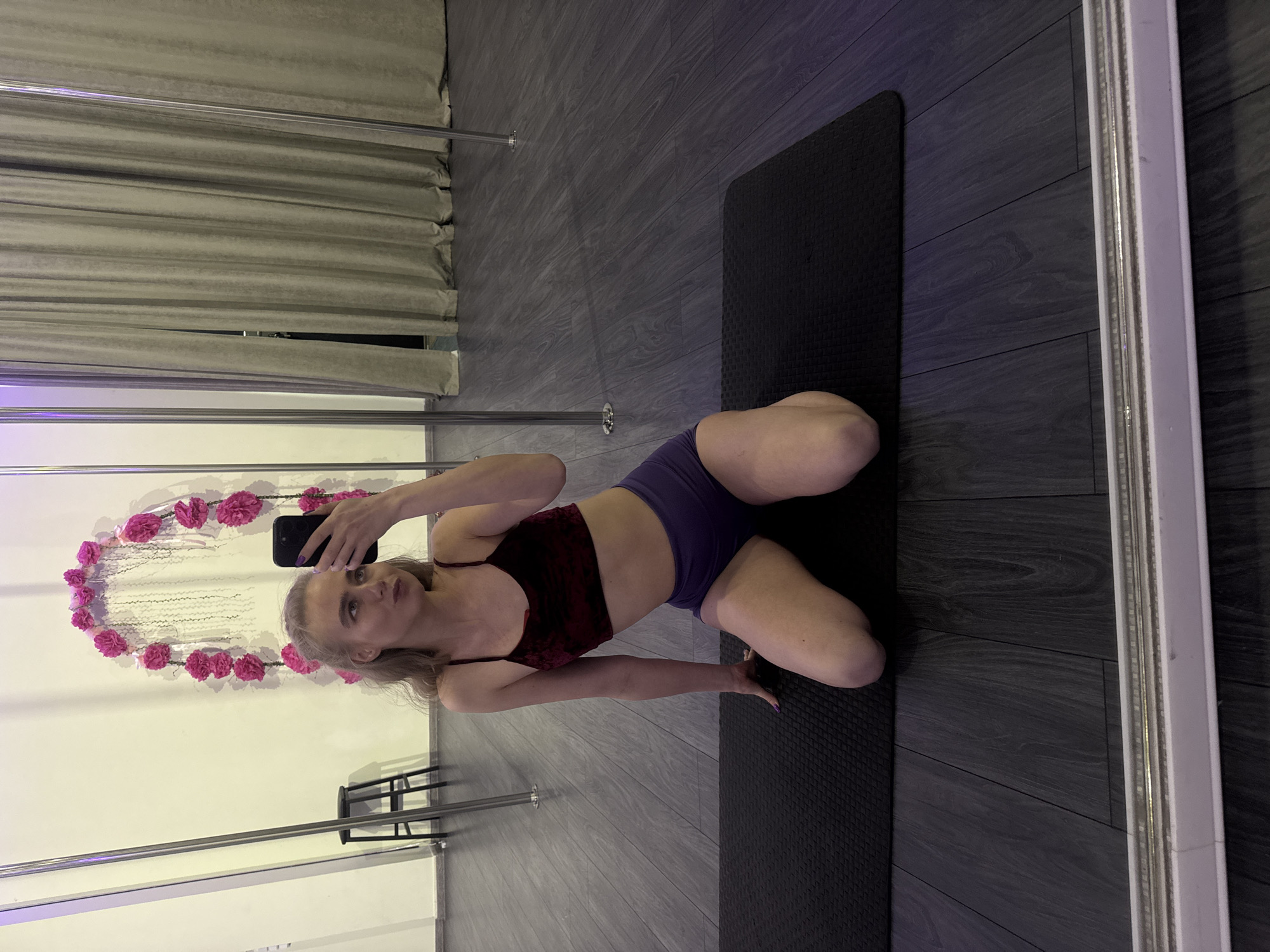 A woman is taking a picture of herself in a mirror. She has blonde hair and is wearing a red velvet top and purple shorts. She is kneeling on a black yoga mat. The background includes a pole with a pink flower wreath, a black stool, grey curtains, and grey wooden flooring. The location is Unknown, Unknown.