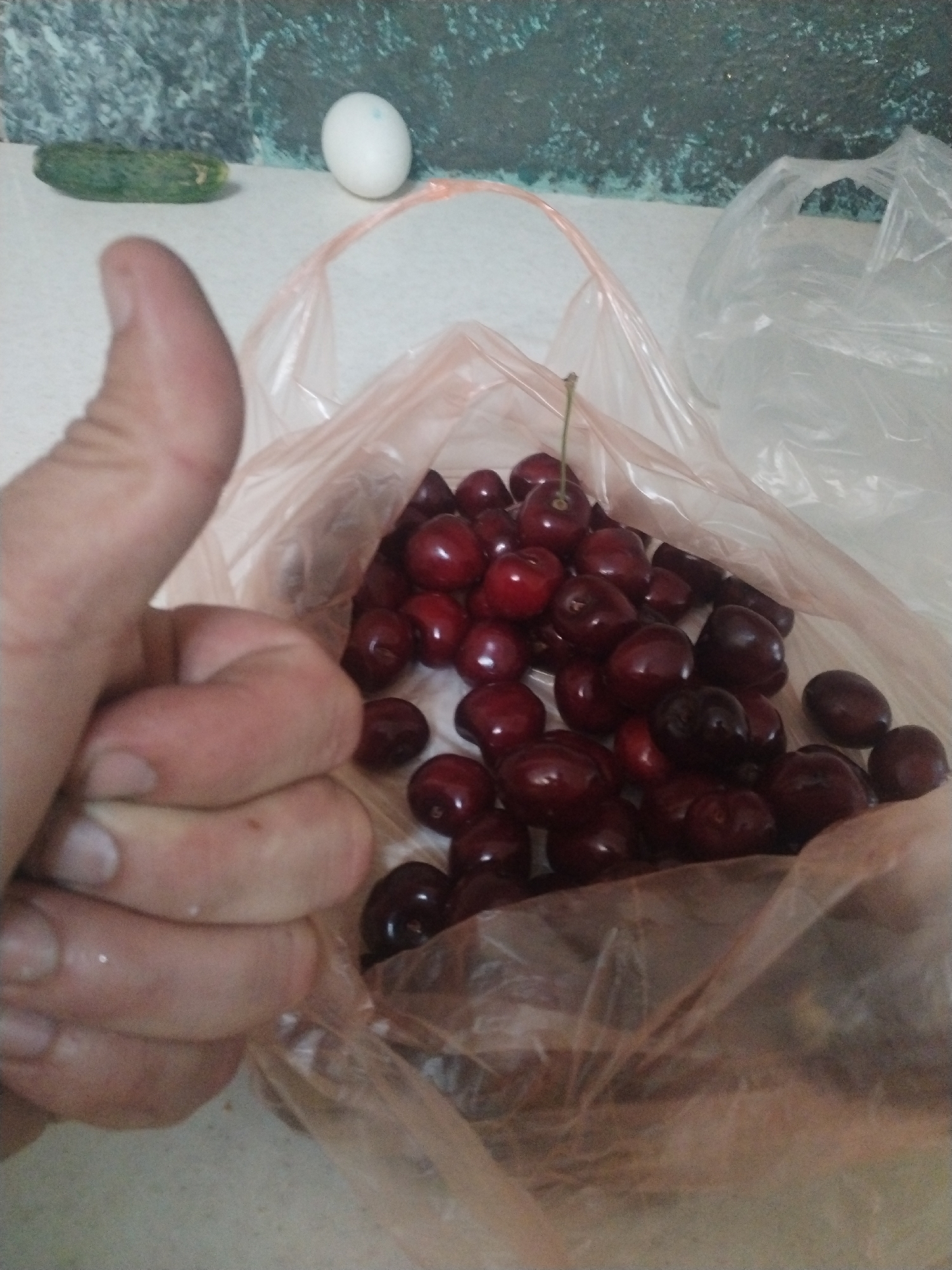 A hand gives a thumbs-up gesture in front of a plastic bag filled with ripe cherries. The bag is transparent and a light pink color, with the cherries spilling out from its opening. A stem is visible on one of the cherries. In the background, a white egg and a cucumber lie on a speckled surface. The setting appears to be a kitchen countertop or similar flat surface. The lighting is somewhat dim, suggesting indoor conditions. The image is a close-up shot, focusing on the hand and the cherries, with the background elements out of focus.