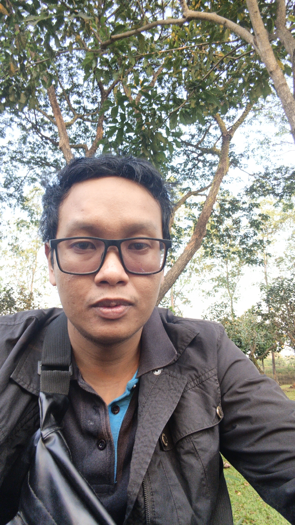A young man wearing glasses and a black jacket with a blue shirt underneath is looking directly at the camera. He has a bag slung across his chest. The background consists of trees and foliage, suggesting an outdoor setting. The image appears to be a selfie taken in Bacin, Indonesia, possibly in a park or natural area.