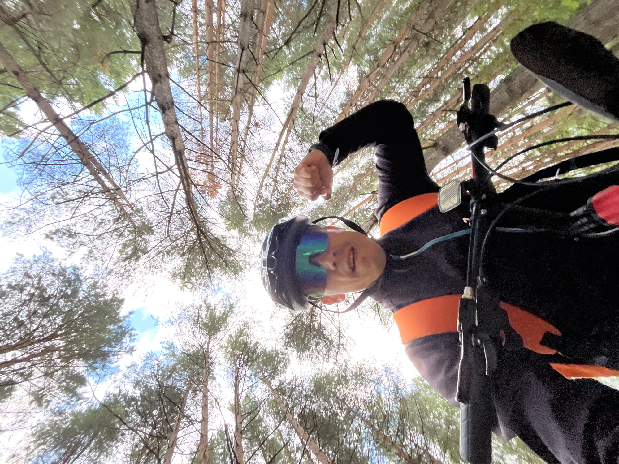 A man wearing a helmet and sunglasses is cycling through a forest. He is looking up at the sky, with his right arm flexed as if to show his muscle. The trees are tall and thin, with their branches reaching towards the sky. The sky is blue with some clouds. The man is wearing a black jacket with an orange vest over it. He is on a bicycle, with the handlebars visible in the foreground. The image appears to be taken from a low angle, looking up at the man and the trees. The location is the city of Chaikovsky, Russia.