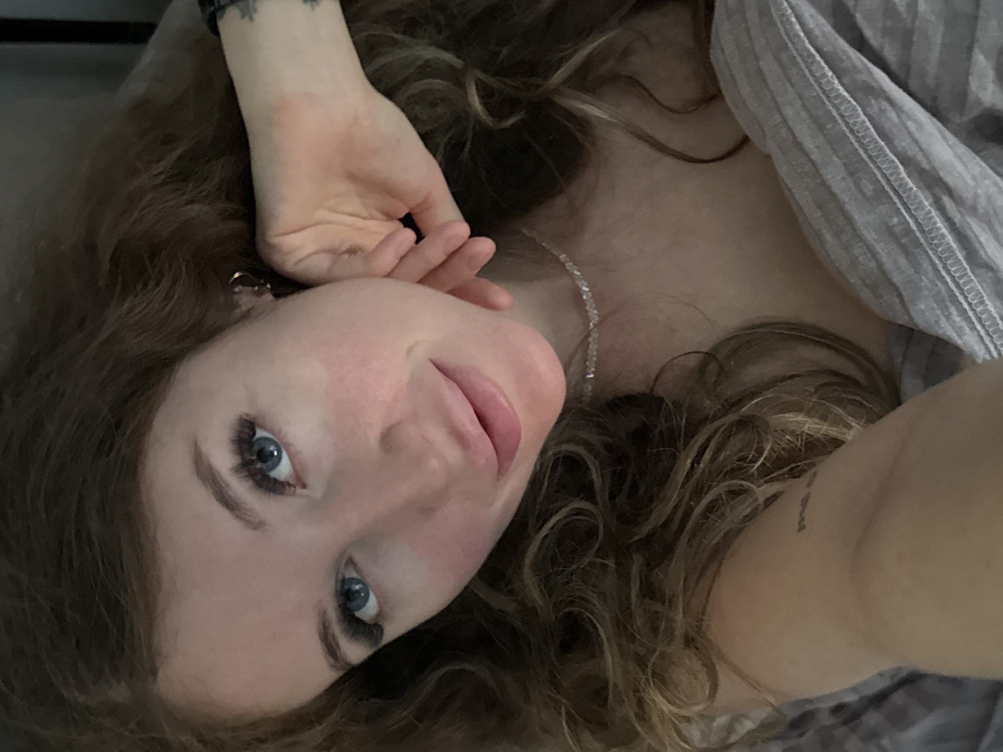 The image is a close-up selfie of a woman lying in bed. She has long, curly brown hair, blue eyes, and light skin with rosy cheeks. She is wearing a delicate, clear beaded necklace and has a tattoo on her forearm. Her right hand is resting near her face, and her left arm is visible at the bottom of the frame, resting on a grey, striped blanket. The lighting suggests it may be indoors, possibly in a bedroom. The image does not contain any recognizable landmarks or features that would indicate a specific location within the city of Chaikovsky, Russia, or any other city.