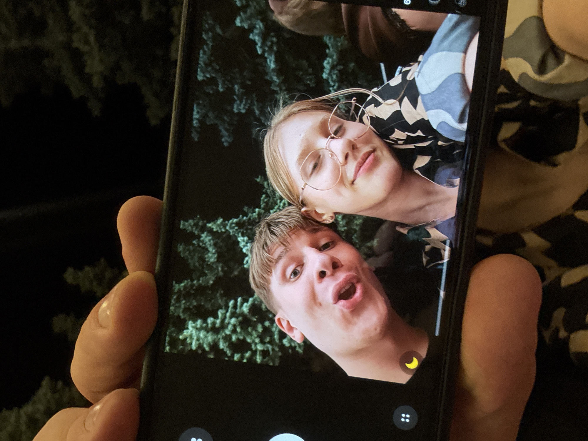A cell phone is held in a person's hand, displaying a selfie of two young adults. The man on the left is making a surprised expression with his mouth open. The woman on the right is wearing glasses and leaning her head on his shoulder. They are both wearing patterned shirts. In the background of the selfie, there are green trees against a dark sky. The setting appears to be outdoors at night. The image does not provide any information about the location in Tiraspol, Moldova.